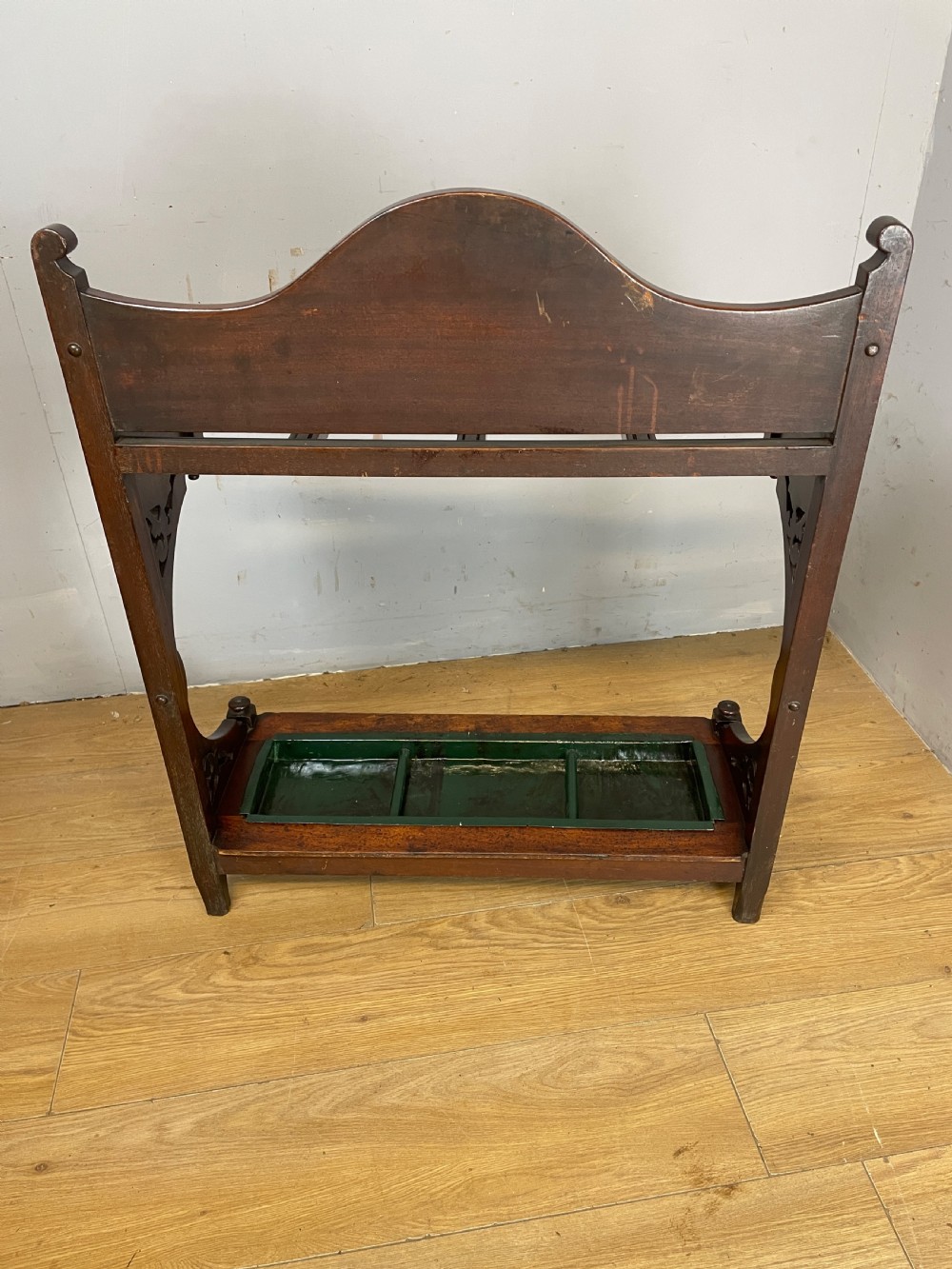 An Arts and Crafts mahogany stick stand featuring a carved central floral motif, divided top rail, turned finials, and a removable green metal drip tray at the base. View VIII