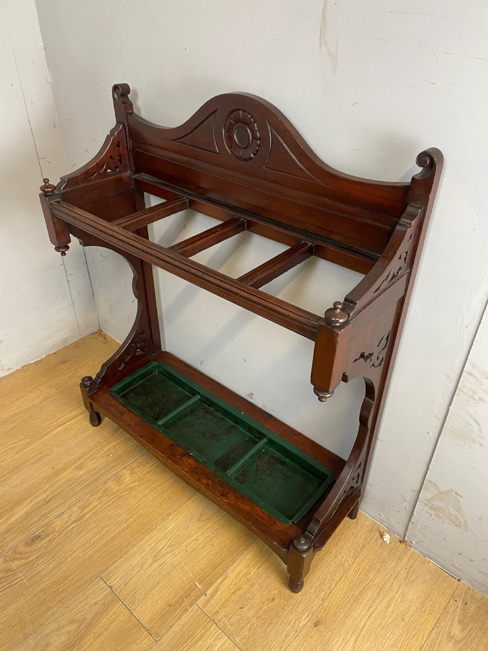 An Arts and Crafts mahogany stick stand featuring a carved central floral motif, divided top rail, turned finials, and a removable green metal drip tray at the base. View IX