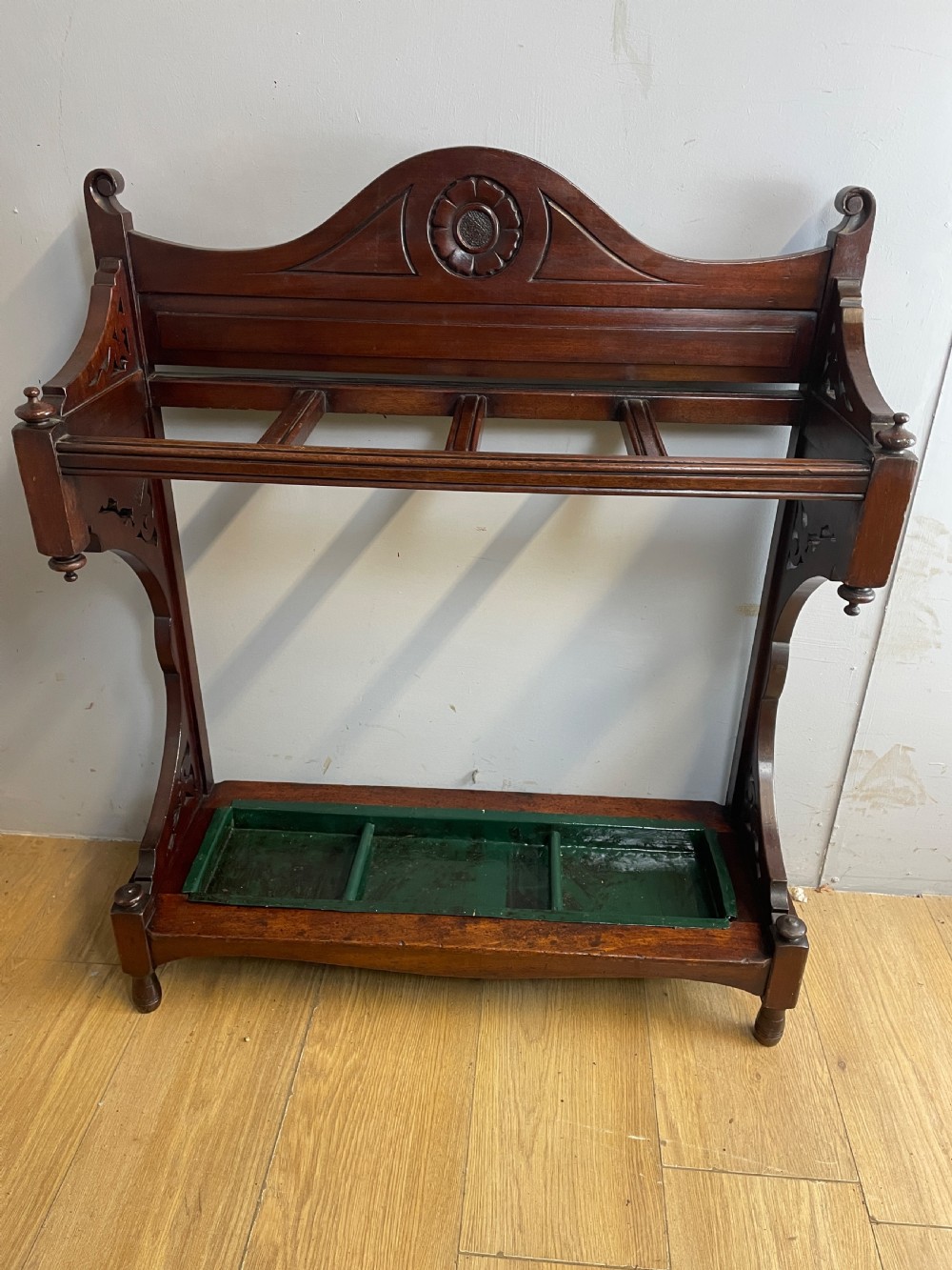 An Arts and Crafts mahogany stick stand featuring a carved central floral motif, divided top rail, turned finials, and a removable green metal drip tray at the base. View X