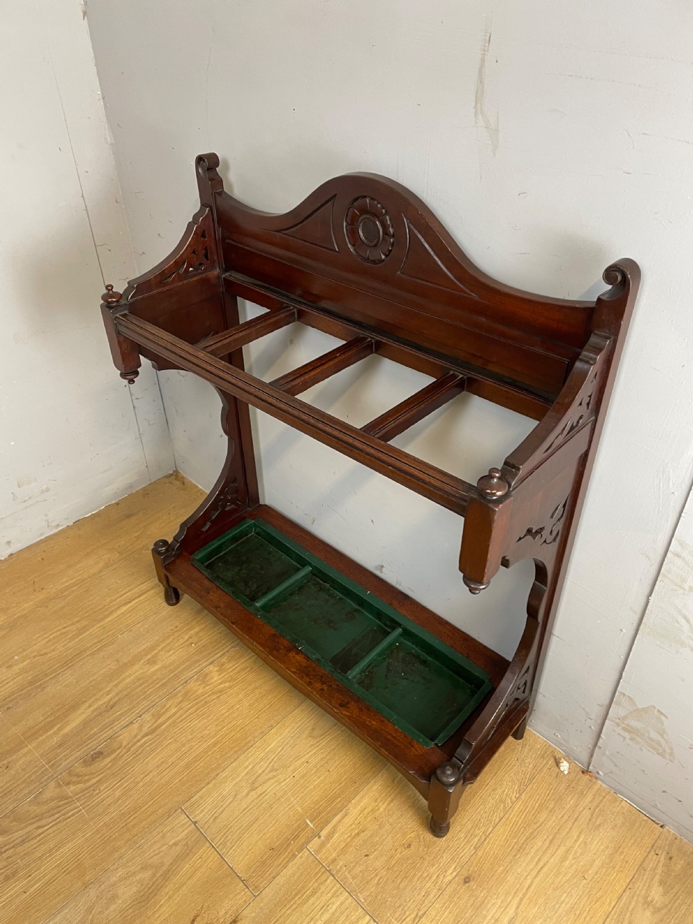 An Arts and Crafts mahogany stick stand featuring a carved central floral motif, divided top rail, turned finials, and a removable green metal drip tray at the base. View XI