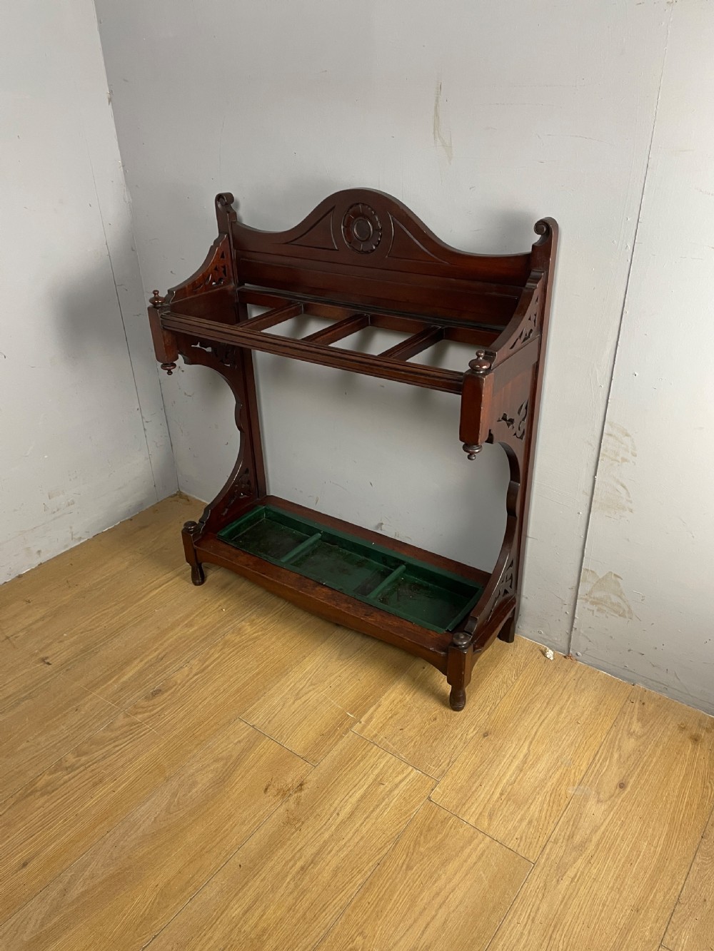 An Arts and Crafts mahogany stick stand featuring a carved central floral motif, divided top rail, turned finials, and a removable green metal drip tray at the base. View XII