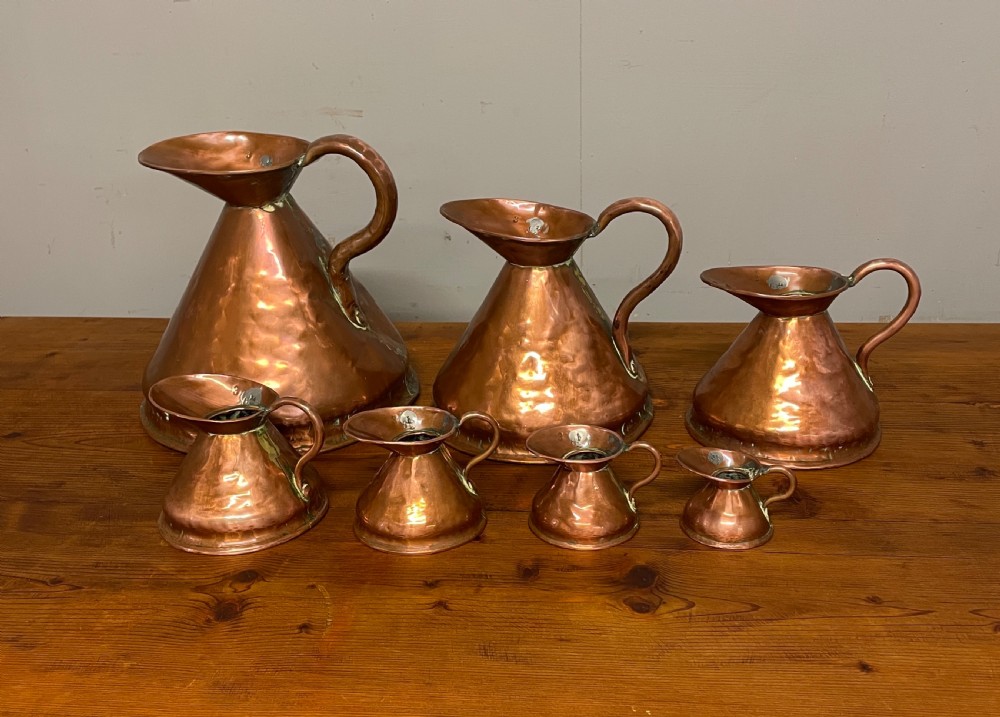 A set of seven graduated Georgian copper measuring jugs featuring haystack bodies and loop handles arranged by size on a wooden surface.