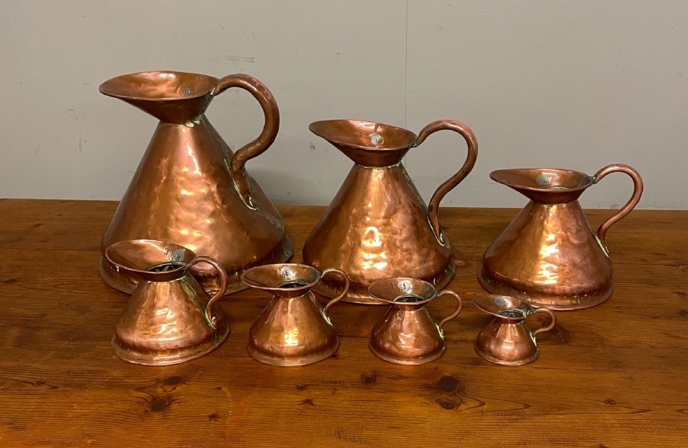 A set of seven graduated Georgian copper measuring jugs featuring haystack bodies and loop handles arranged by size on a wooden surface. View II