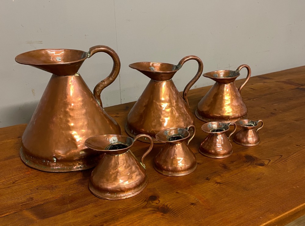 A set of seven graduated Georgian copper measuring jugs featuring haystack bodies and loop handles arranged by size on a wooden surface. View III