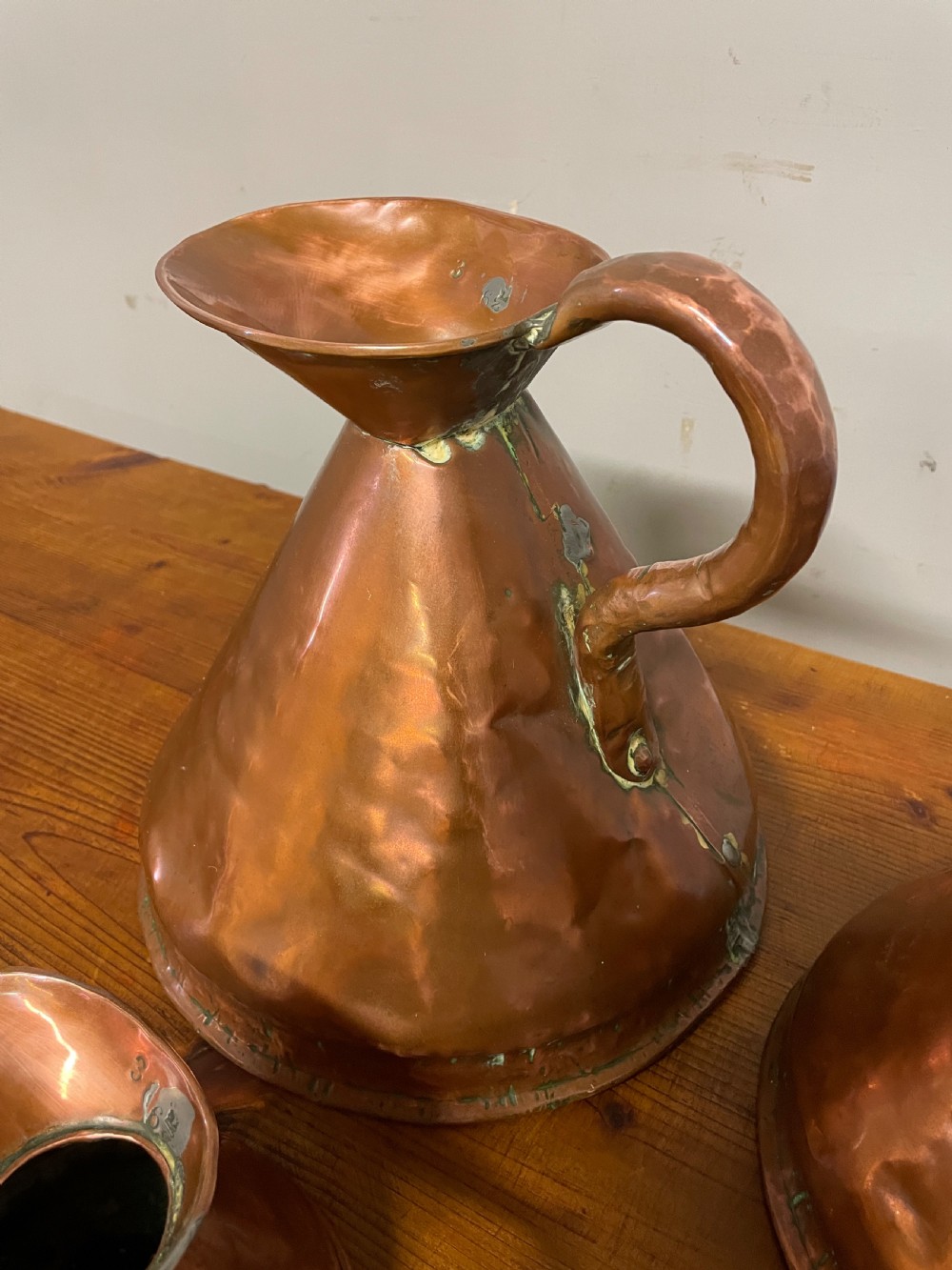 A set of seven graduated Georgian copper measuring jugs featuring haystack bodies and loop handles arranged by size on a wooden surface. View IV
