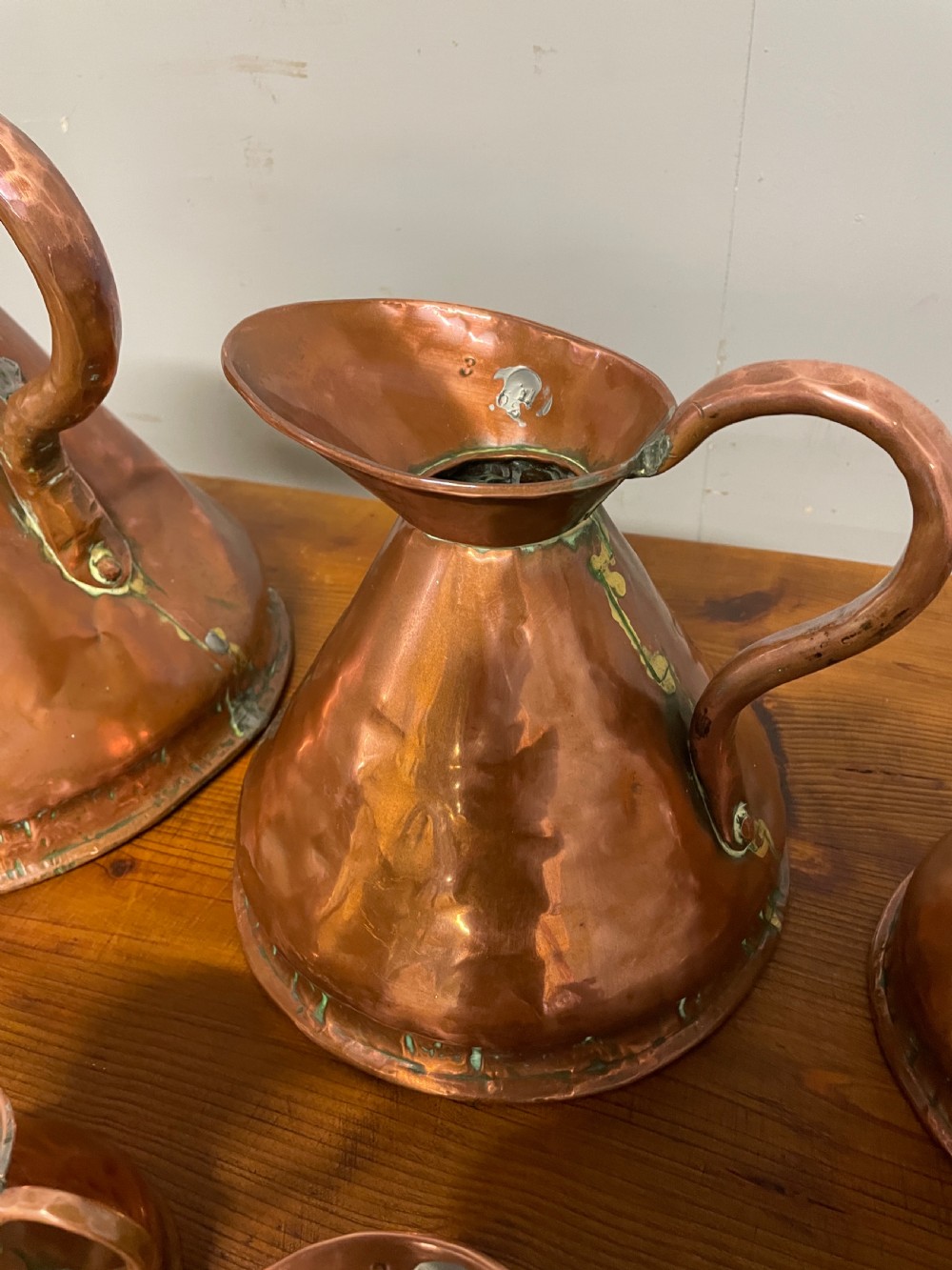 A set of seven graduated Georgian copper measuring jugs featuring haystack bodies and loop handles arranged by size on a wooden surface. View V