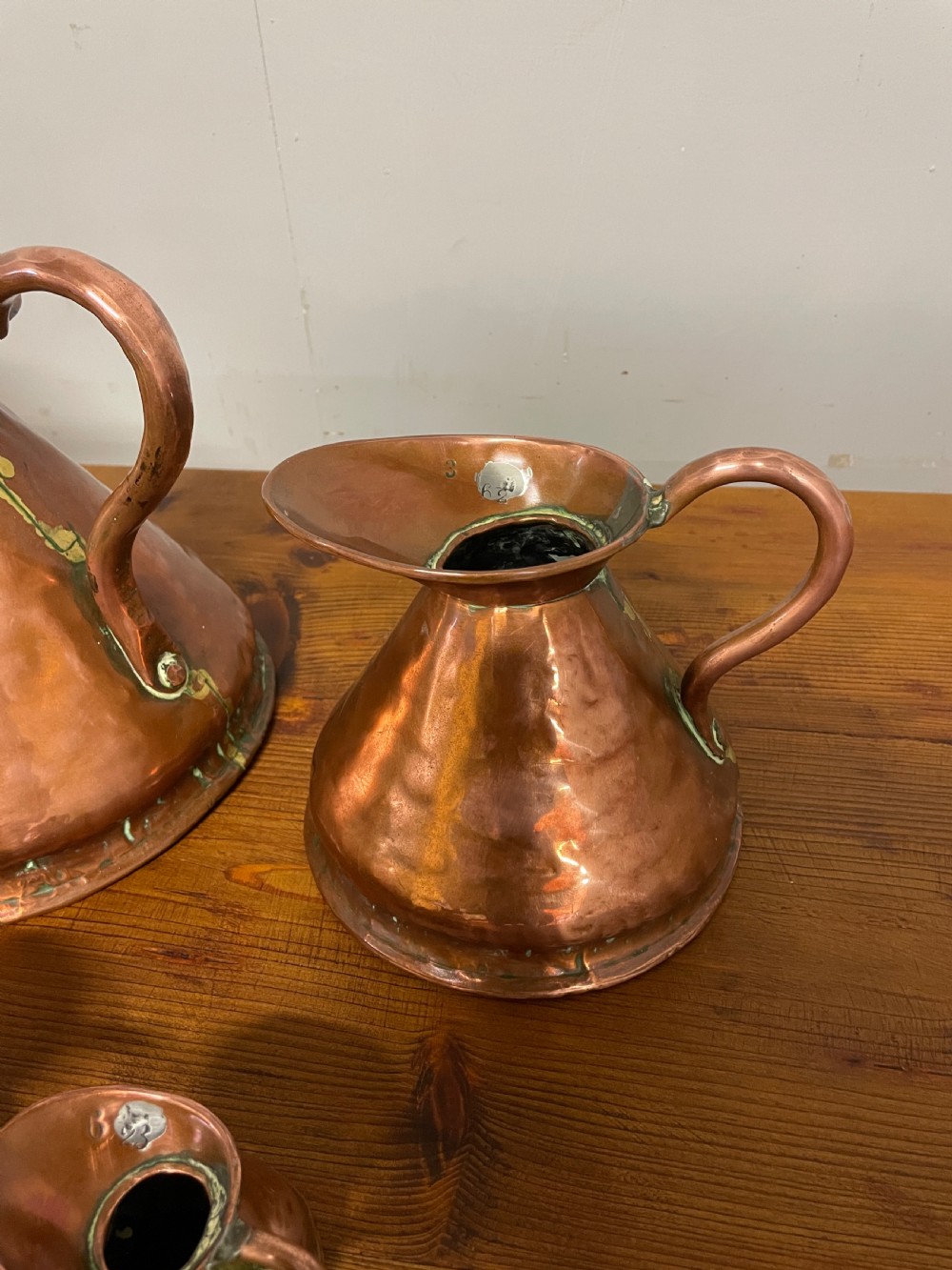 A set of seven graduated Georgian copper measuring jugs featuring haystack bodies and loop handles arranged by size on a wooden surface. View VI