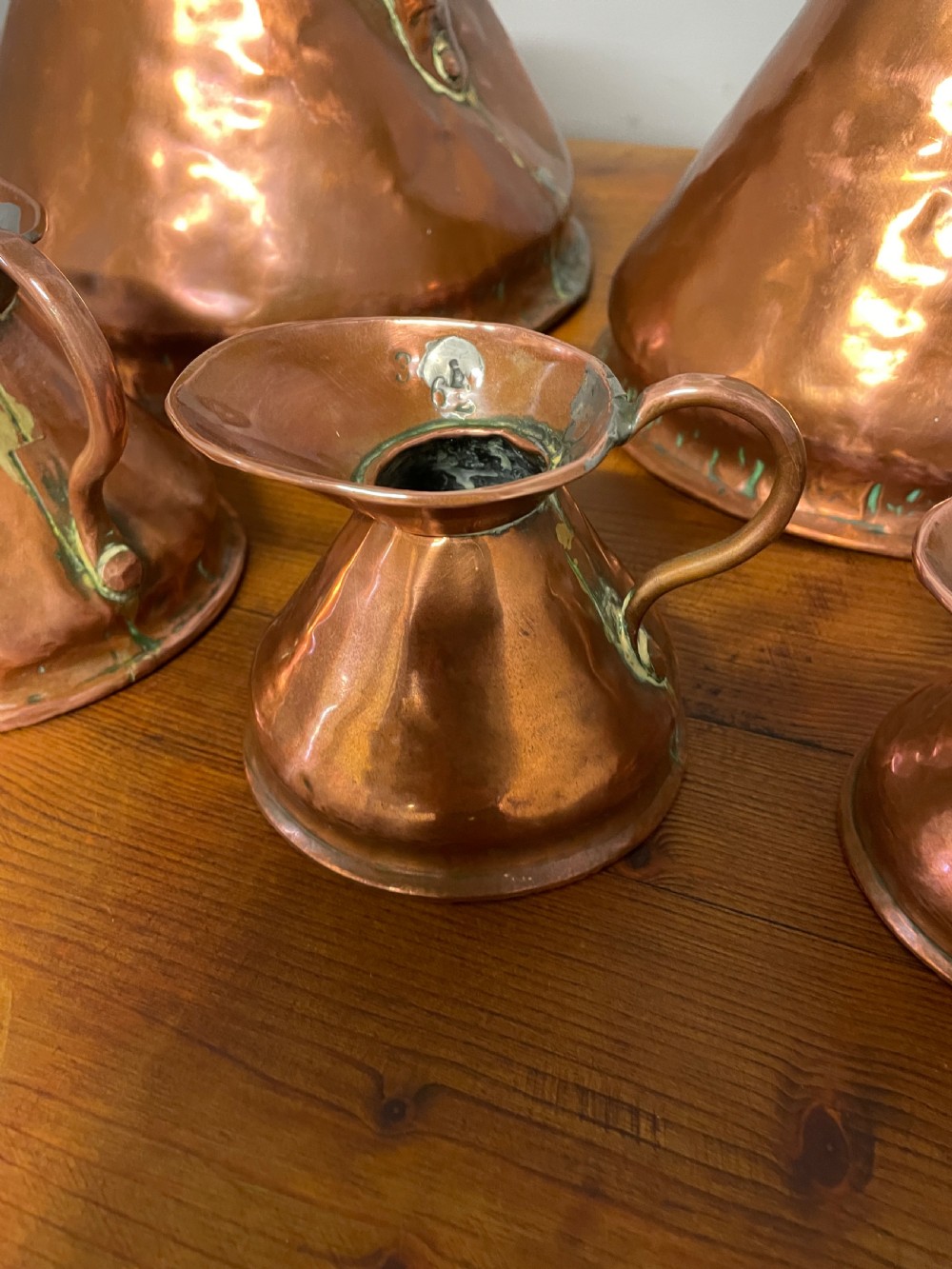 A set of seven graduated Georgian copper measuring jugs featuring haystack bodies and loop handles arranged by size on a wooden surface. View VIII
