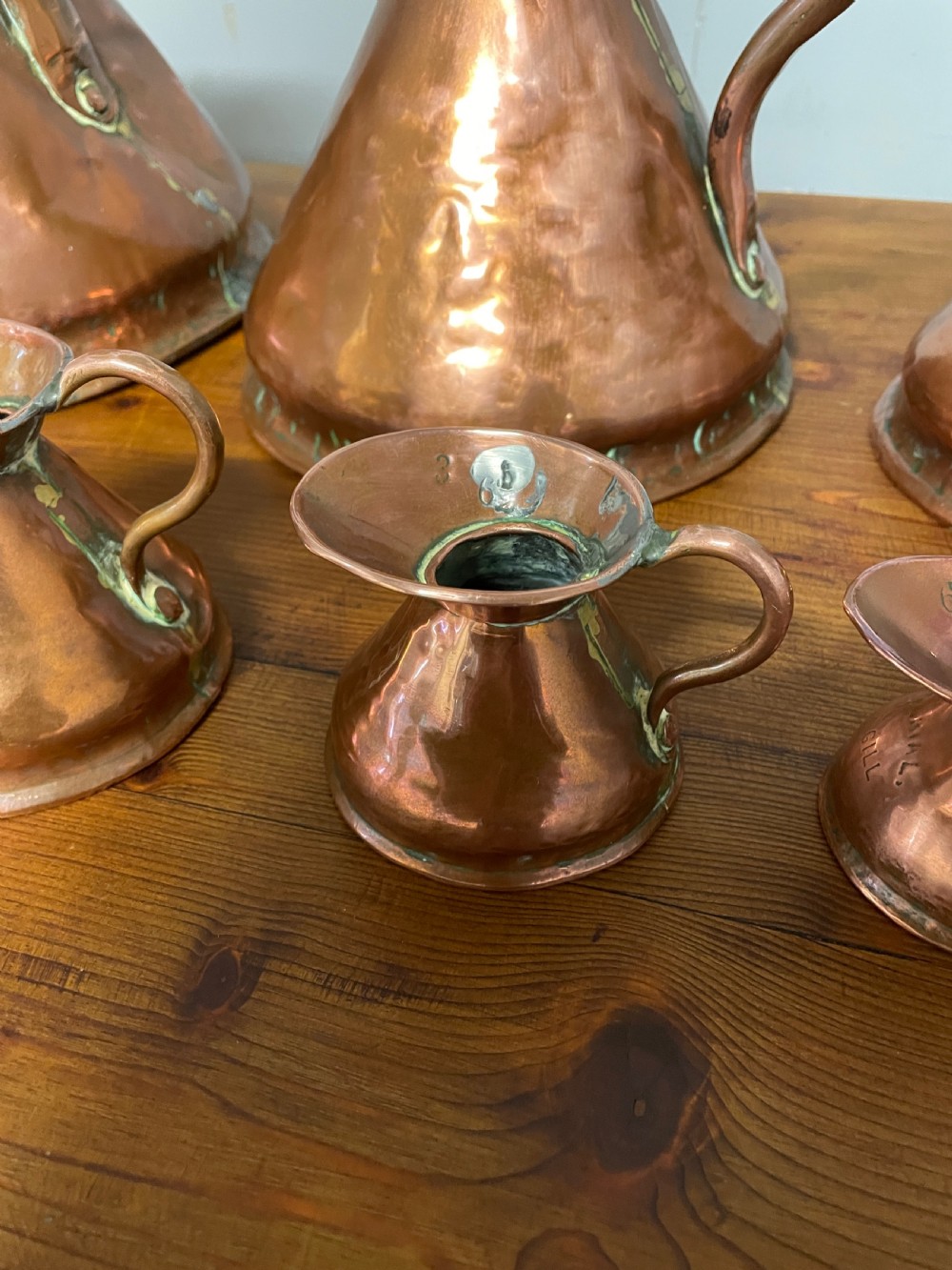 A set of seven graduated Georgian copper measuring jugs featuring haystack bodies and loop handles arranged by size on a wooden surface. View IX