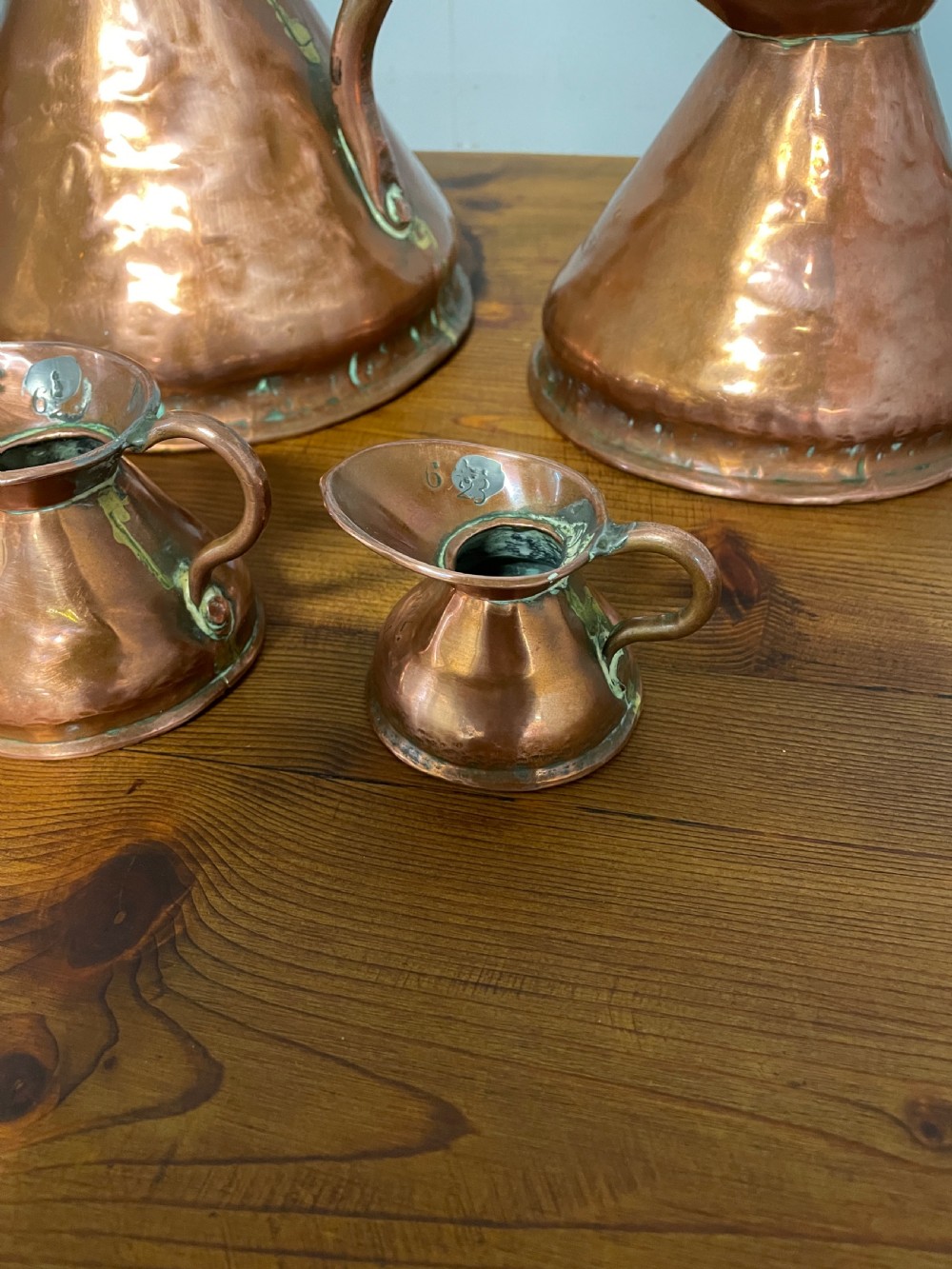 A set of seven graduated Georgian copper measuring jugs featuring haystack bodies and loop handles arranged by size on a wooden surface. View X