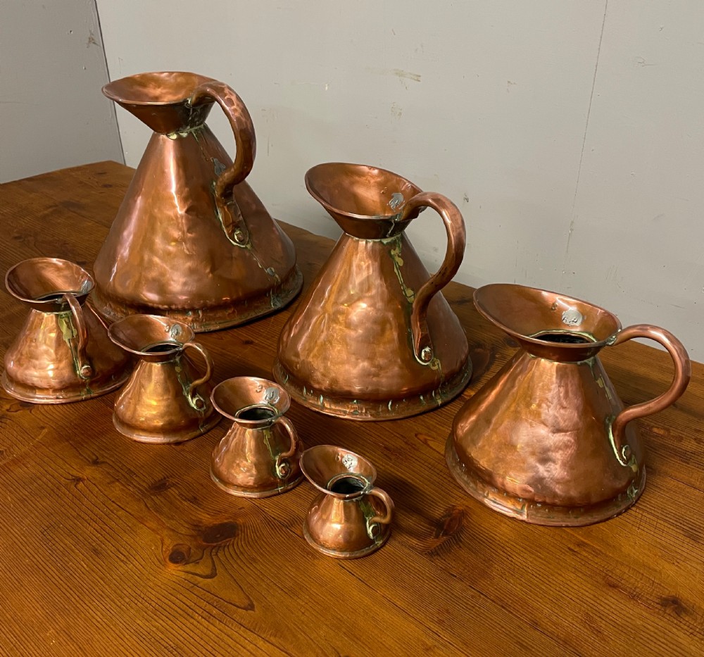 A set of seven graduated Georgian copper measuring jugs featuring haystack bodies and loop handles arranged by size on a wooden surface. View XVII