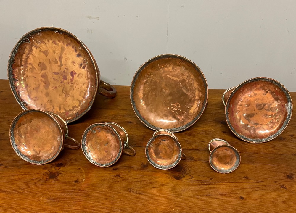 A set of seven graduated Georgian copper measuring jugs featuring haystack bodies and loop handles arranged by size on a wooden surface. View XVIII