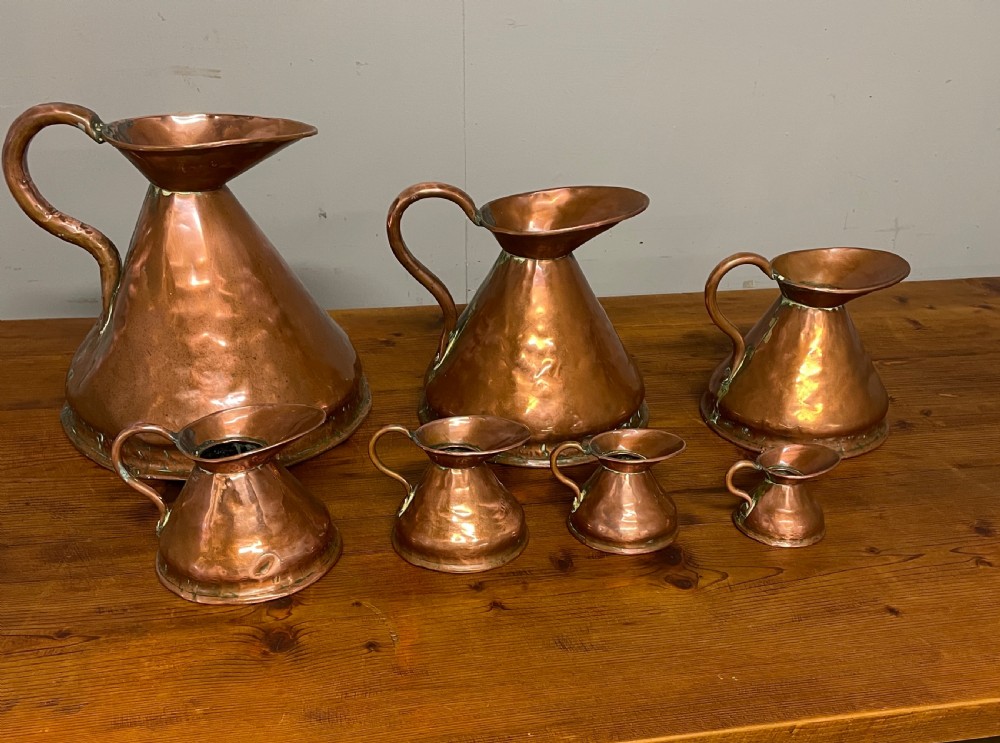 A set of seven graduated Georgian copper measuring jugs featuring haystack bodies and loop handles arranged by size on a wooden surface. View XXI