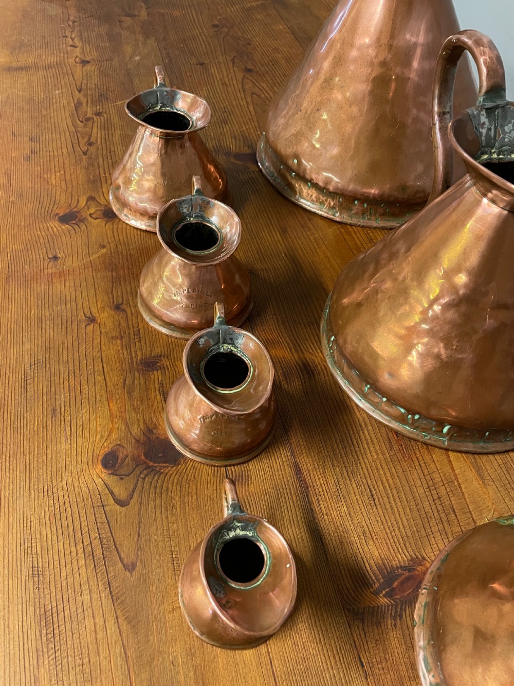 A set of seven graduated Georgian copper measuring jugs featuring haystack bodies and loop handles arranged by size on a wooden surface. View XXIV
