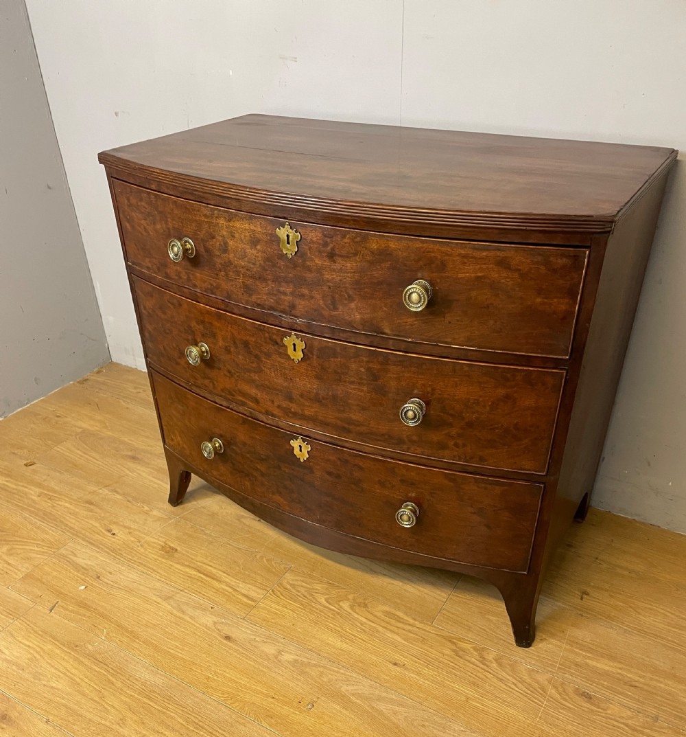 An antique Regency mahogany bow-front chest of drawers featuring three long drawers with brass knobs, brass keyhole escutcheons, and curved bracket feet.