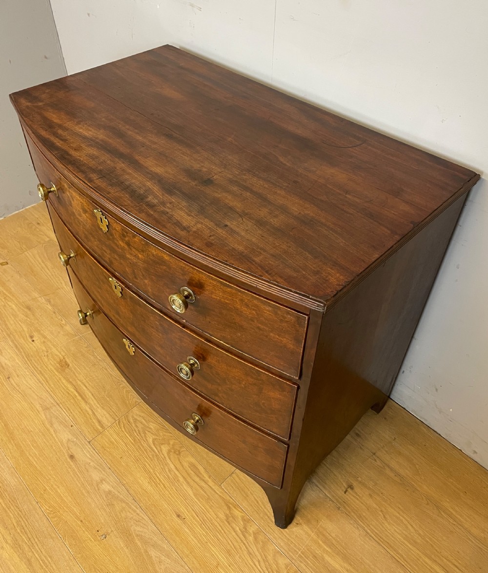 An antique Regency mahogany bow-front chest of drawers featuring three long drawers with brass knobs, brass keyhole escutcheons, and curved bracket feet. View VI