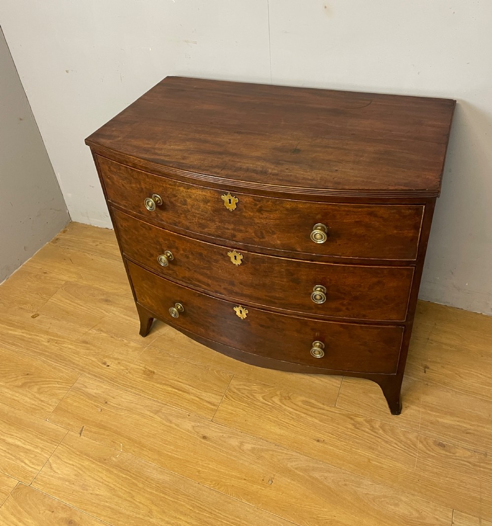 An antique Regency mahogany bow-front chest of drawers featuring three long drawers with brass knobs, brass keyhole escutcheons, and curved bracket feet. View IX