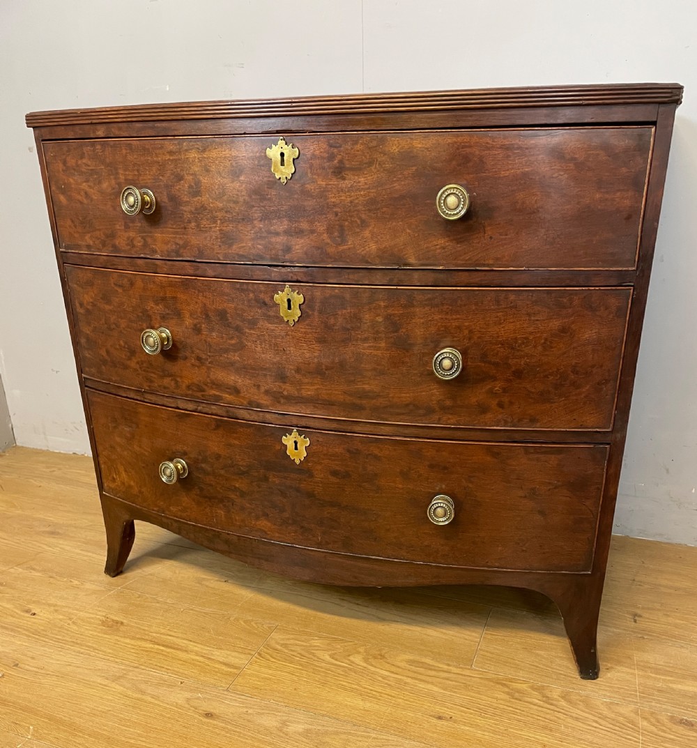 An antique Regency mahogany bow-front chest of drawers featuring three long drawers with brass knobs, brass keyhole escutcheons, and curved bracket feet. View X