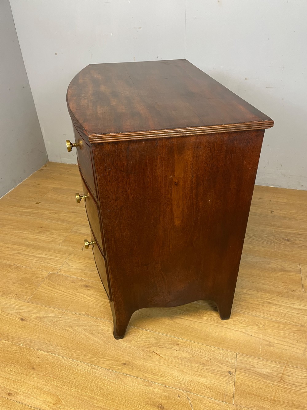 An antique Regency mahogany bow-front chest of drawers featuring three long drawers with brass knobs, brass keyhole escutcheons, and curved bracket feet. View XI
