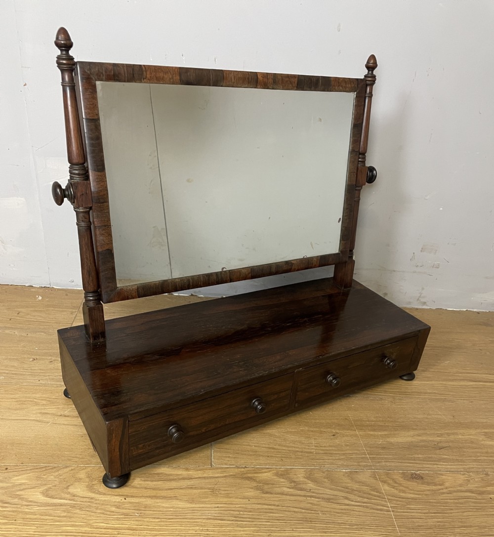 An early Victorian dark rosewood dressing table mirror featuring turned support columns, finials, and a rectangular swivelling frame resting on a plinth drawer base.