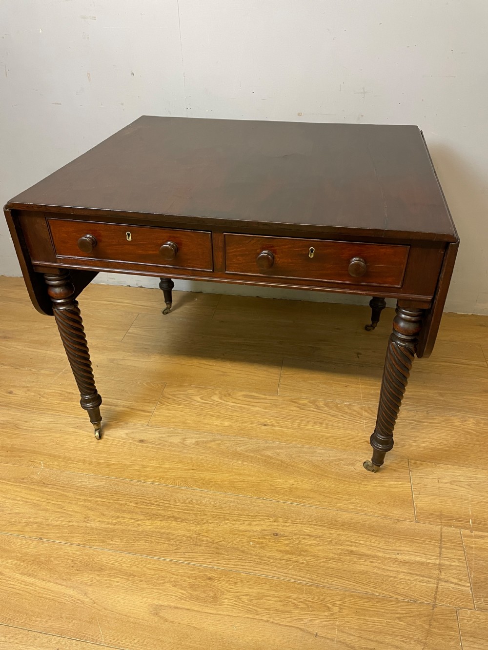 A dark mahogany Regency writing table featuring two drawers with wooden knobs, drop-leaf sides, and spiral-twisted legs on brass castors.