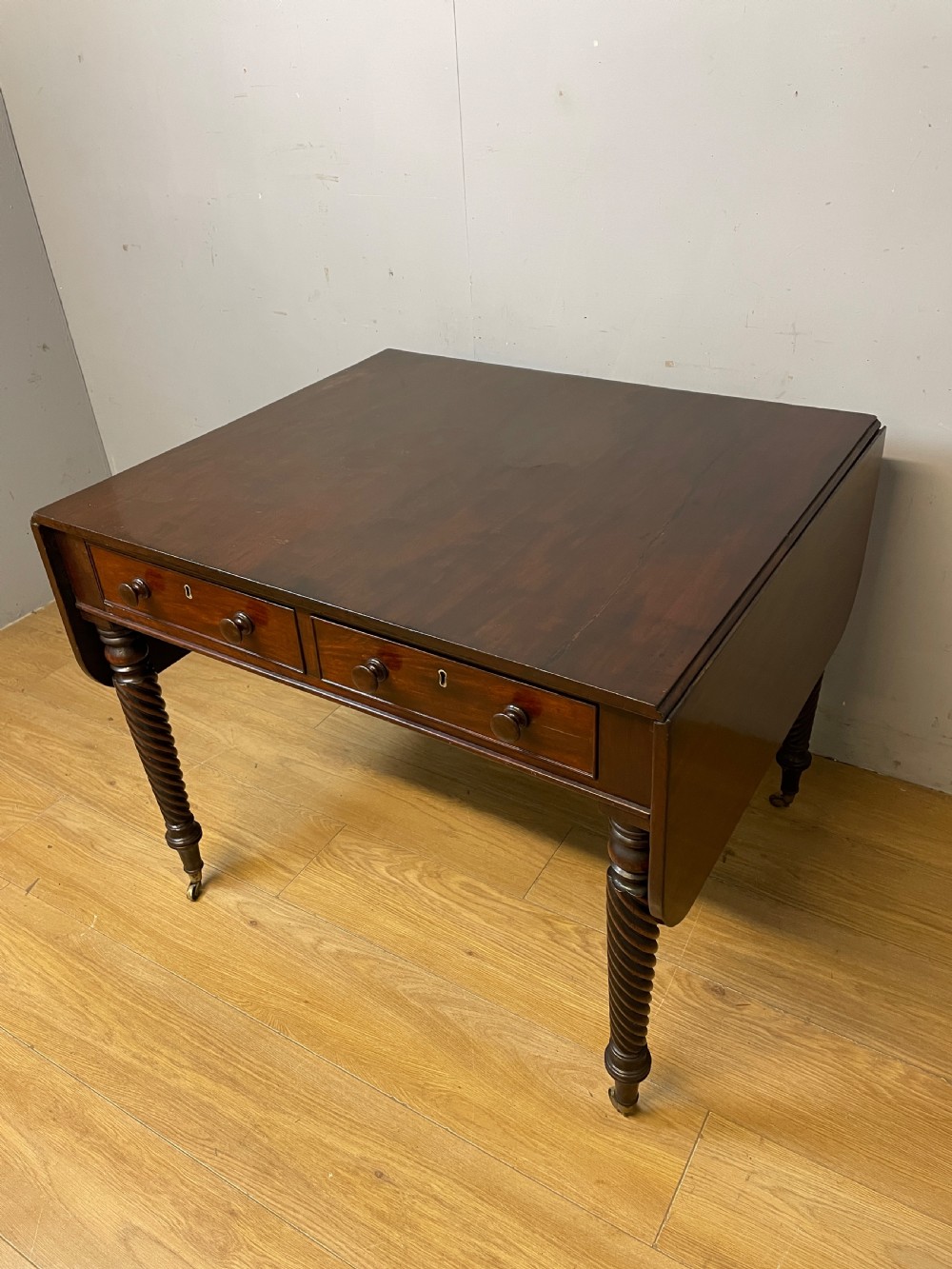 A dark mahogany Regency writing table featuring two drawers with wooden knobs, drop-leaf sides, and spiral-twisted legs on brass castors. View II