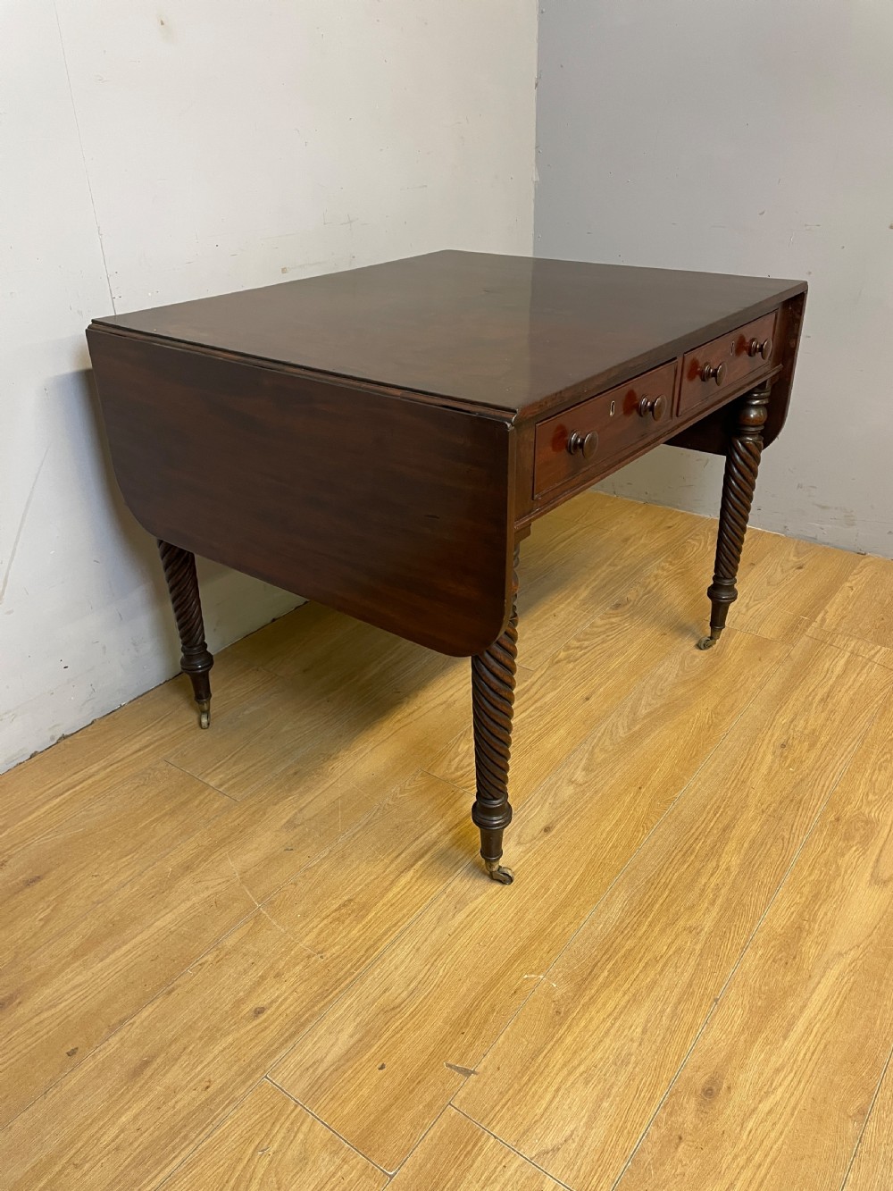 A dark mahogany Regency writing table featuring two drawers with wooden knobs, drop-leaf sides, and spiral-twisted legs on brass castors. View IV