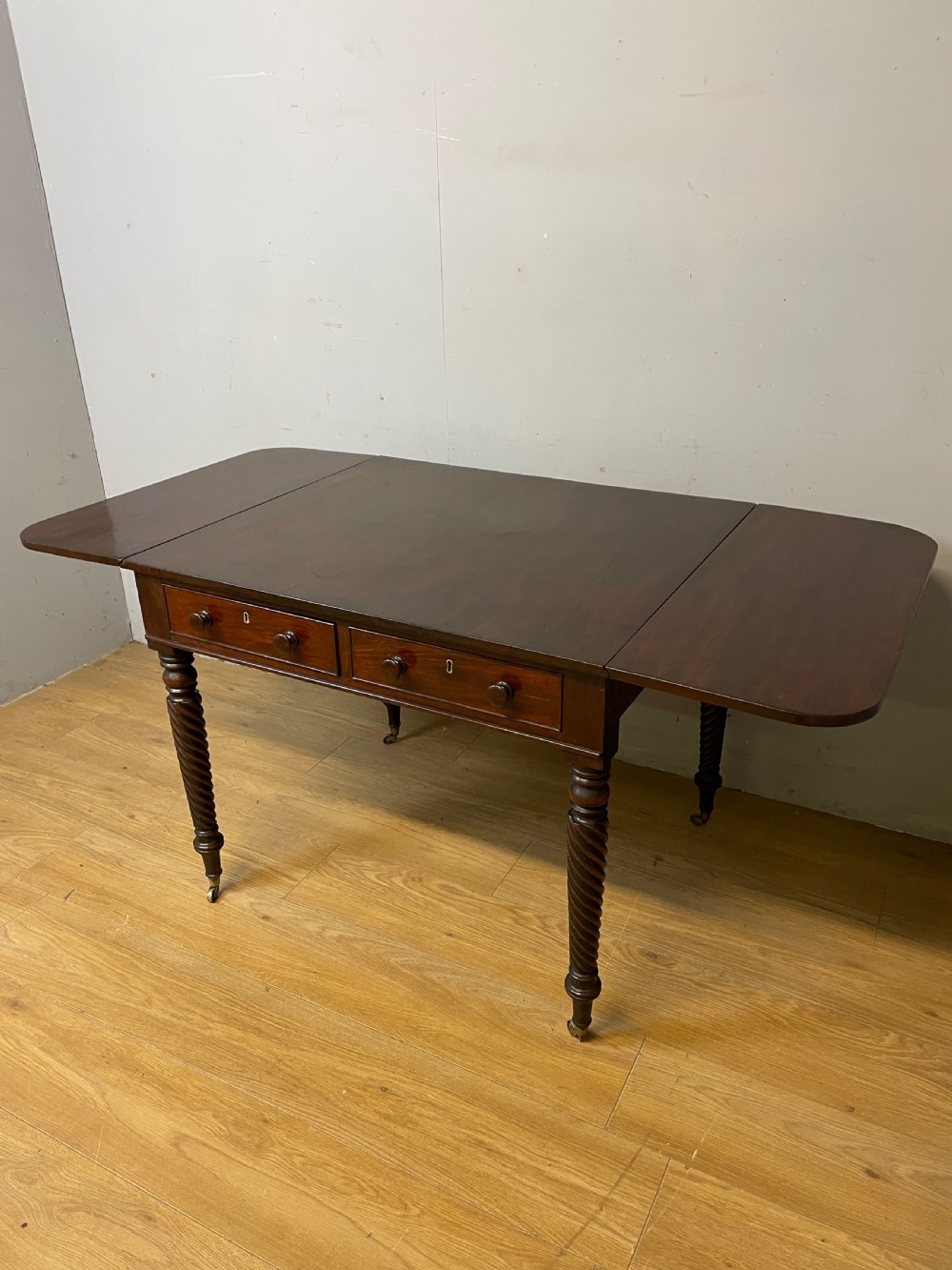 A dark mahogany Regency writing table featuring two drawers with wooden knobs, drop-leaf sides, and spiral-twisted legs on brass castors. View VI