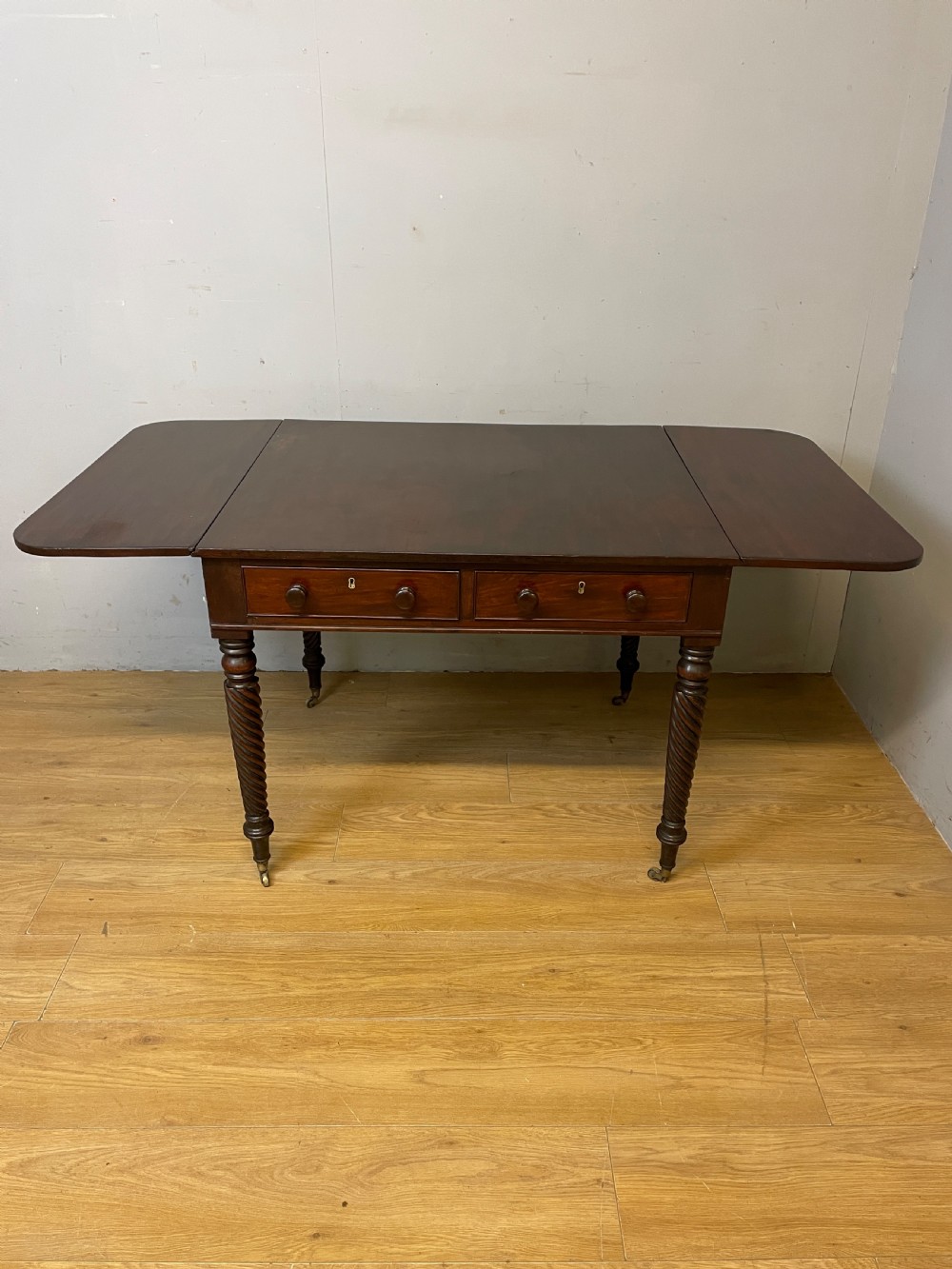 A dark mahogany Regency writing table featuring two drawers with wooden knobs, drop-leaf sides, and spiral-twisted legs on brass castors. View VII