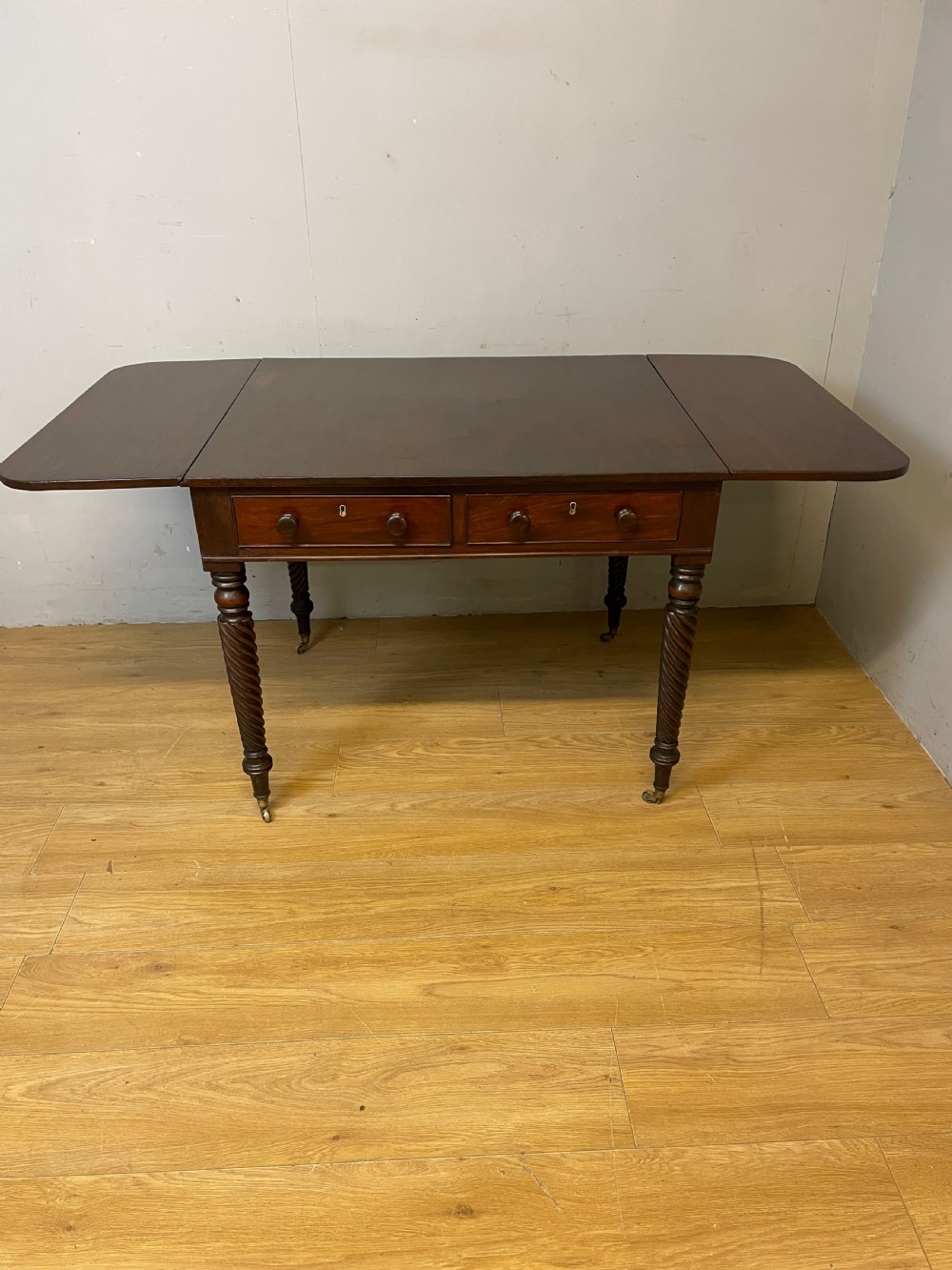 A dark mahogany Regency writing table featuring two drawers with wooden knobs, drop-leaf sides, and spiral-twisted legs on brass castors. View X