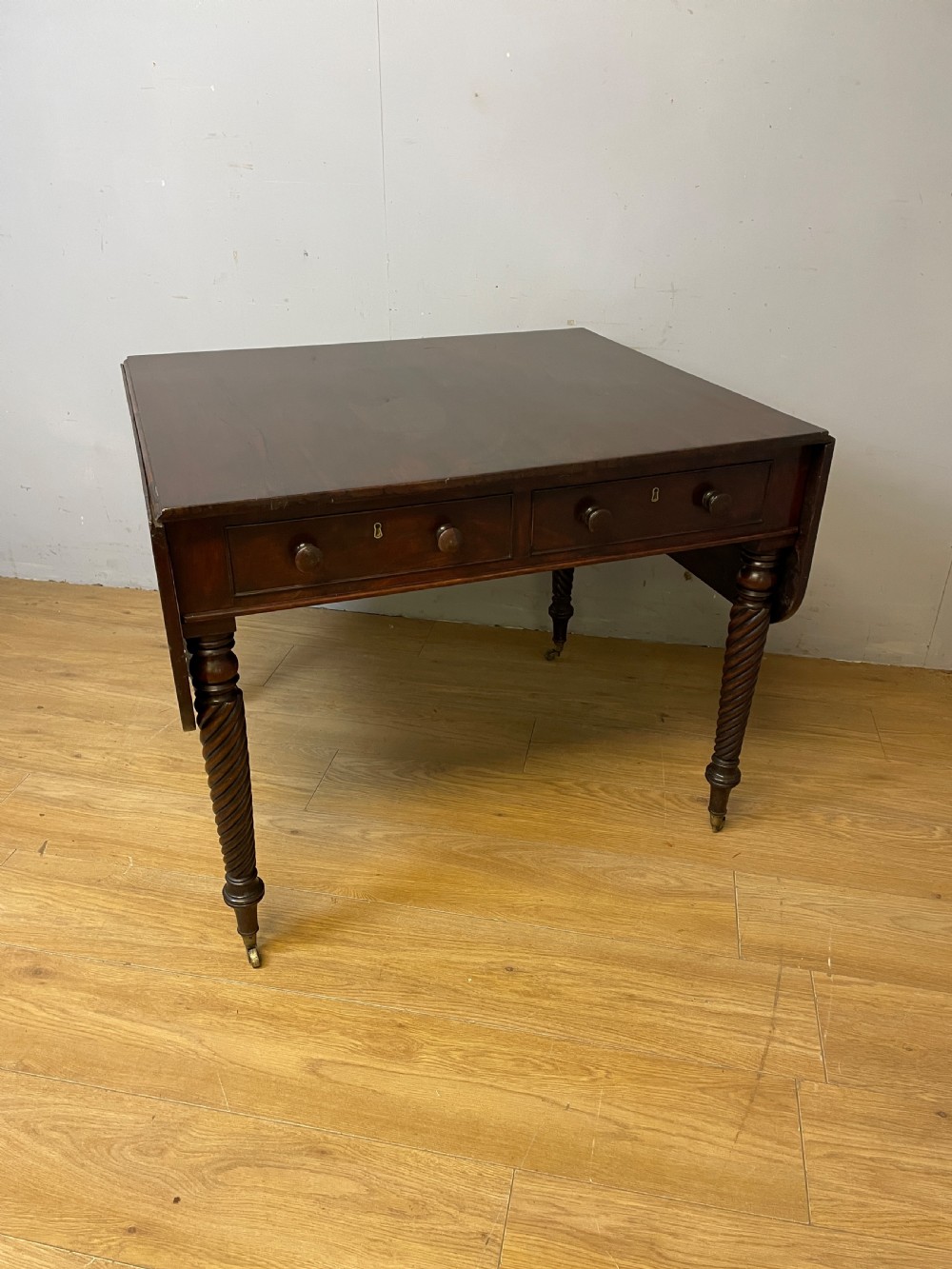 A dark mahogany Regency writing table featuring two drawers with wooden knobs, drop-leaf sides, and spiral-twisted legs on brass castors. View XI