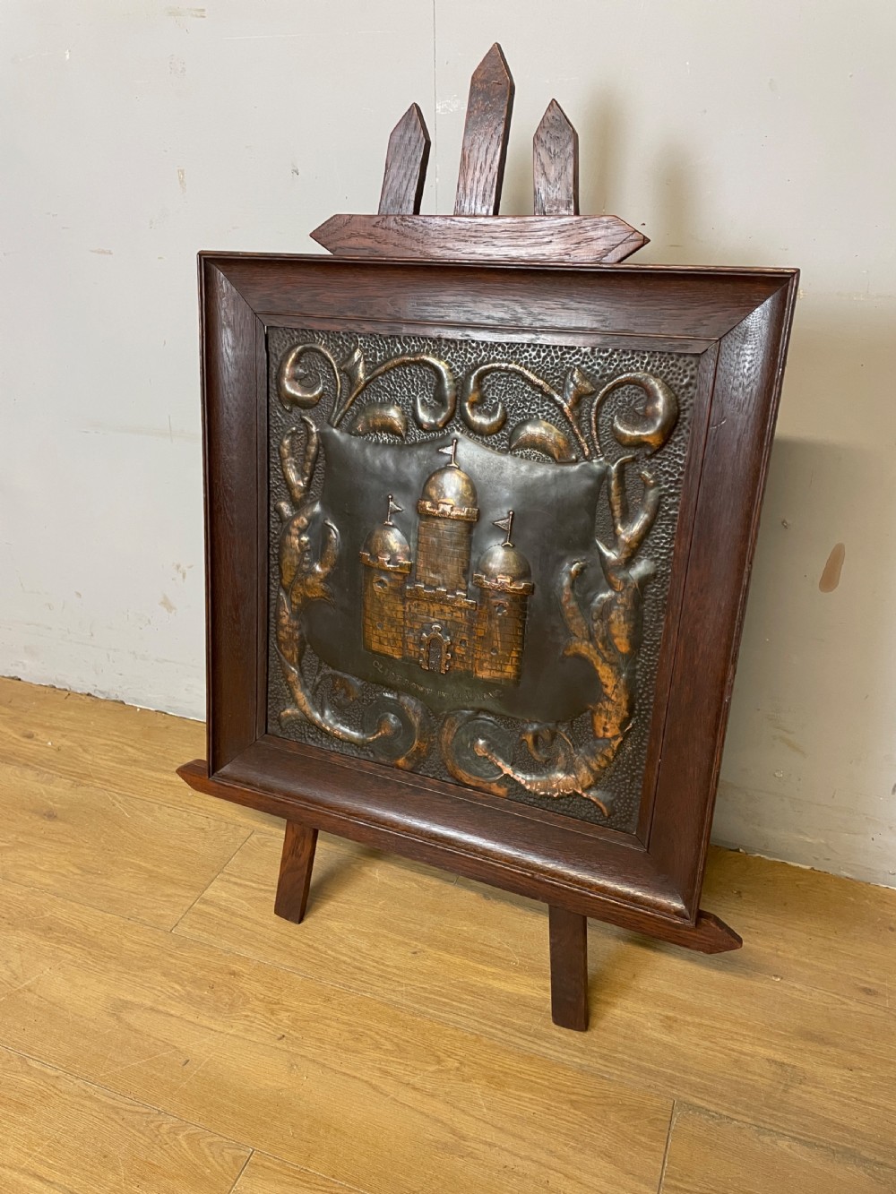 An Arts and Crafts dark oak framed firescreen featuring a central hand-hammered copper panel with an embossed castle motif and scrollwork.