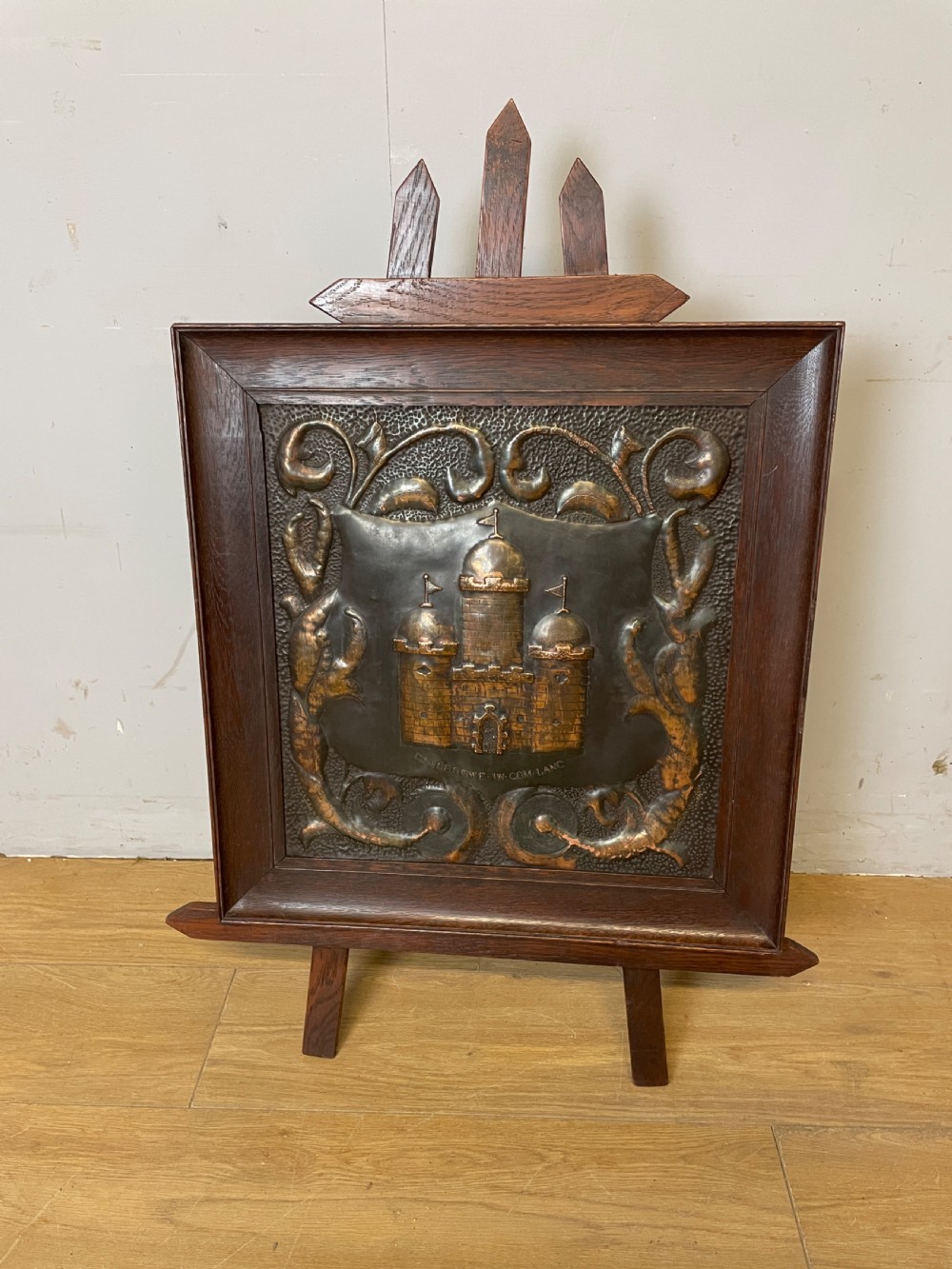 An Arts and Crafts dark oak framed firescreen featuring a central hand-hammered copper panel with an embossed castle motif and scrollwork. View II