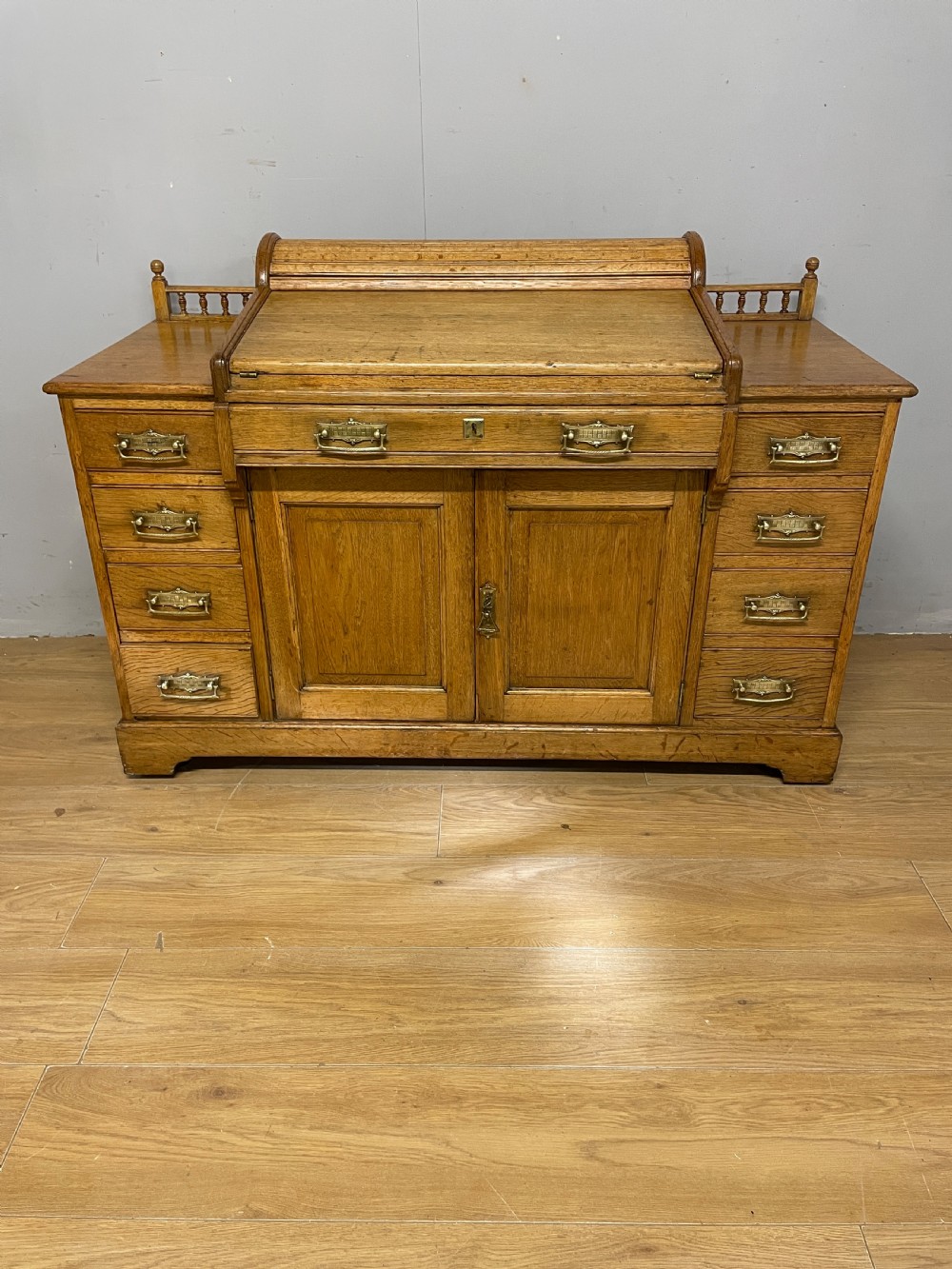 A nineteenth-century golden oak tambour desk featuring multiple side drawers with brass handles, central panelled cupboard doors, and a raised wooden gallery rail.