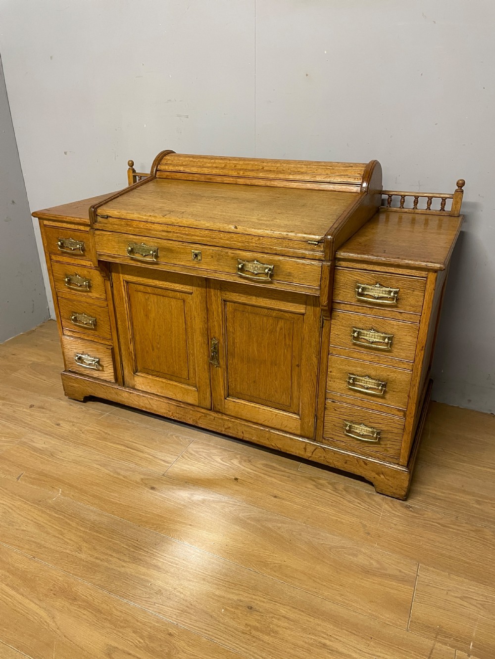 A nineteenth-century golden oak tambour desk featuring multiple side drawers with brass handles, central panelled cupboard doors, and a raised wooden gallery rail. View II