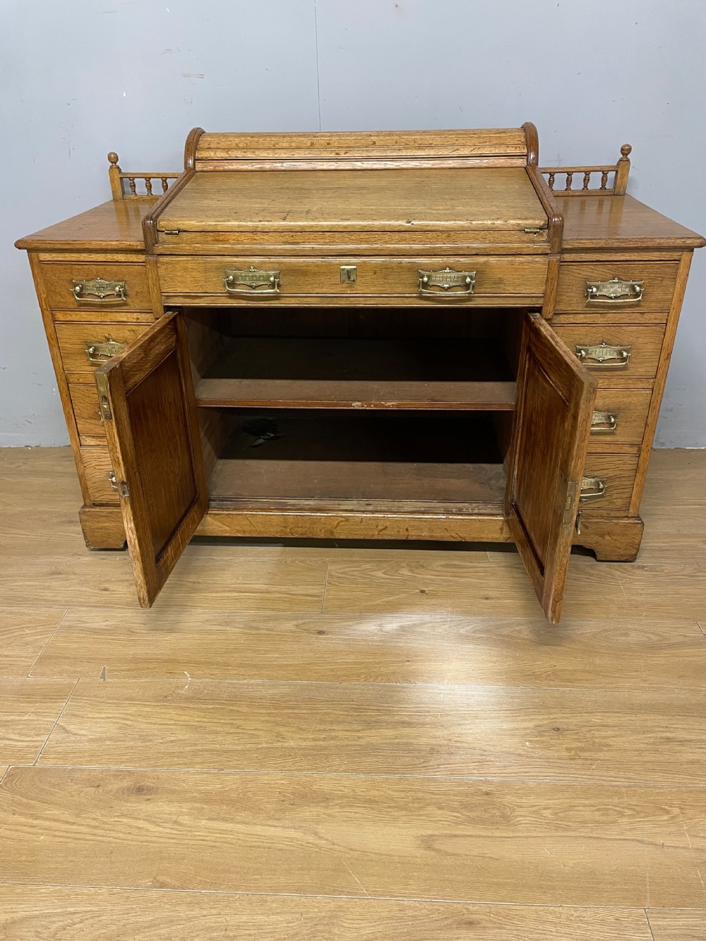 A nineteenth-century golden oak tambour desk featuring multiple side drawers with brass handles, central panelled cupboard doors, and a raised wooden gallery rail. View VII