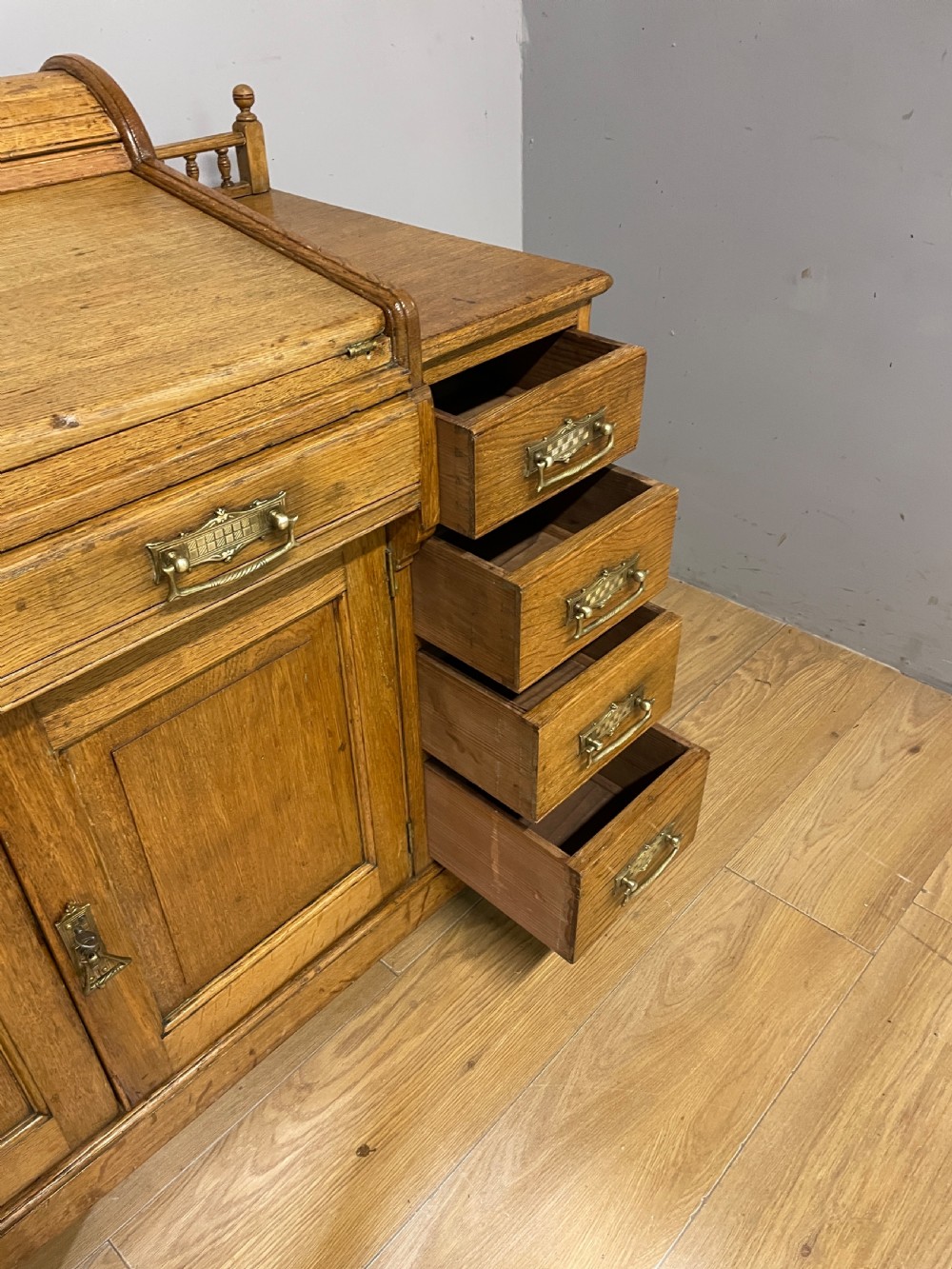 A nineteenth-century golden oak tambour desk featuring multiple side drawers with brass handles, central panelled cupboard doors, and a raised wooden gallery rail. View VIII
