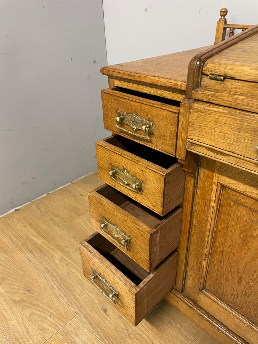 A nineteenth-century golden oak tambour desk featuring multiple side drawers with brass handles, central panelled cupboard doors, and a raised wooden gallery rail. View IX