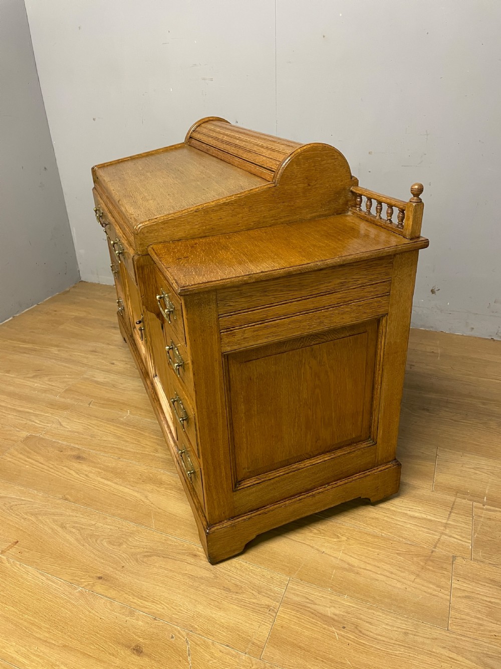 A nineteenth-century golden oak tambour desk featuring multiple side drawers with brass handles, central panelled cupboard doors, and a raised wooden gallery rail. View X