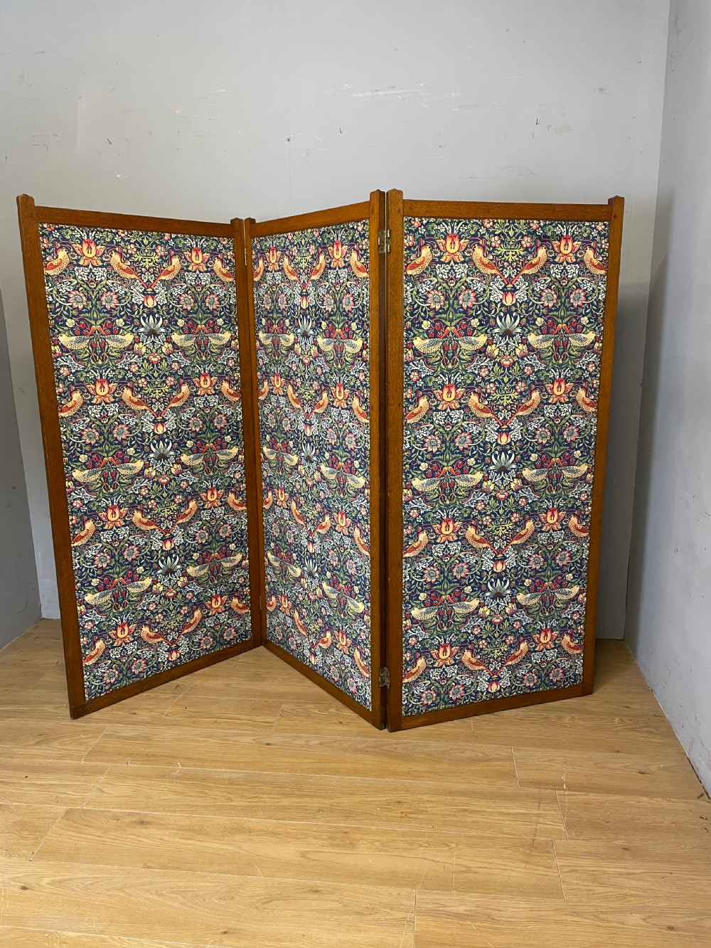 A three-panel wooden folding room screen framed in medium oak and upholstered with William Morris Strawberry Thief pattern velvet fabric in blue, red, and yellow tones.