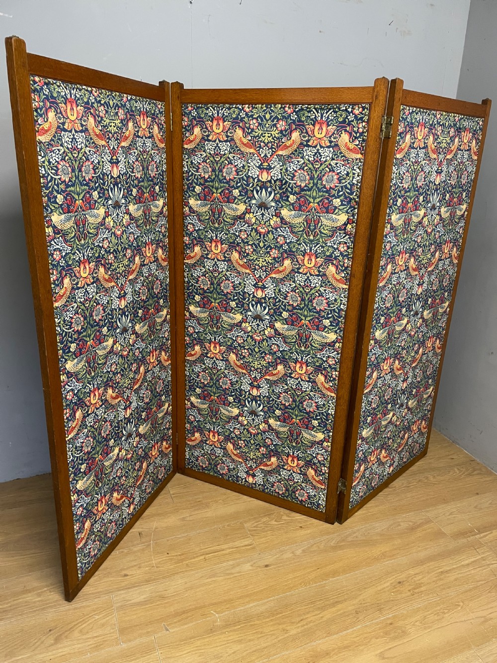 A three-panel wooden folding room screen framed in medium oak and upholstered with William Morris Strawberry Thief pattern velvet fabric in blue, red, and yellow tones. View V