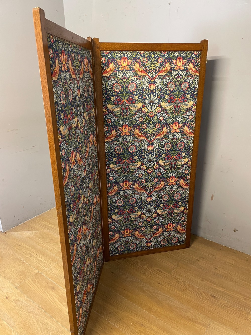 A three-panel wooden folding room screen framed in medium oak and upholstered with William Morris Strawberry Thief pattern velvet fabric in blue, red, and yellow tones. View VII
