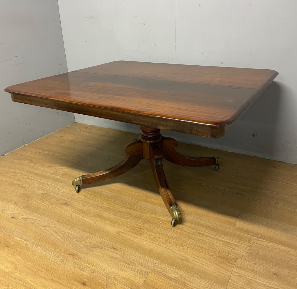 A William IV mahogany tilt-top dining table featuring a rectangular polished top with rounded corners supported by a central turned pedestal and four splayed legs with brass castors.