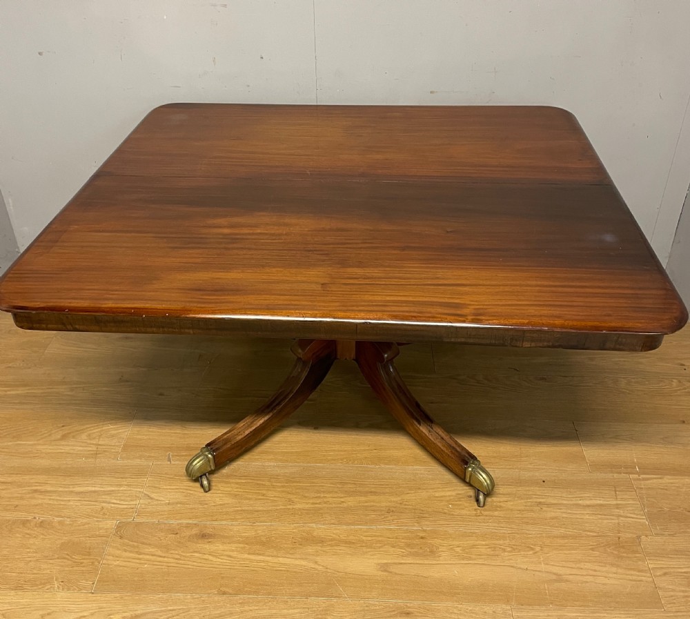 A William IV mahogany tilt-top dining table featuring a rectangular polished top with rounded corners supported by a central turned pedestal and four splayed legs with brass castors. View II