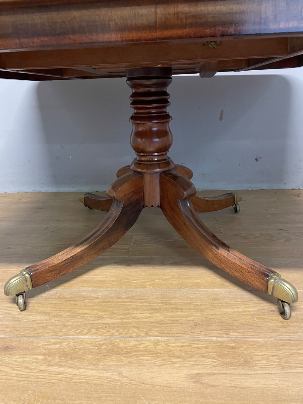 A William IV mahogany tilt-top dining table featuring a rectangular polished top with rounded corners supported by a central turned pedestal and four splayed legs with brass castors. View III