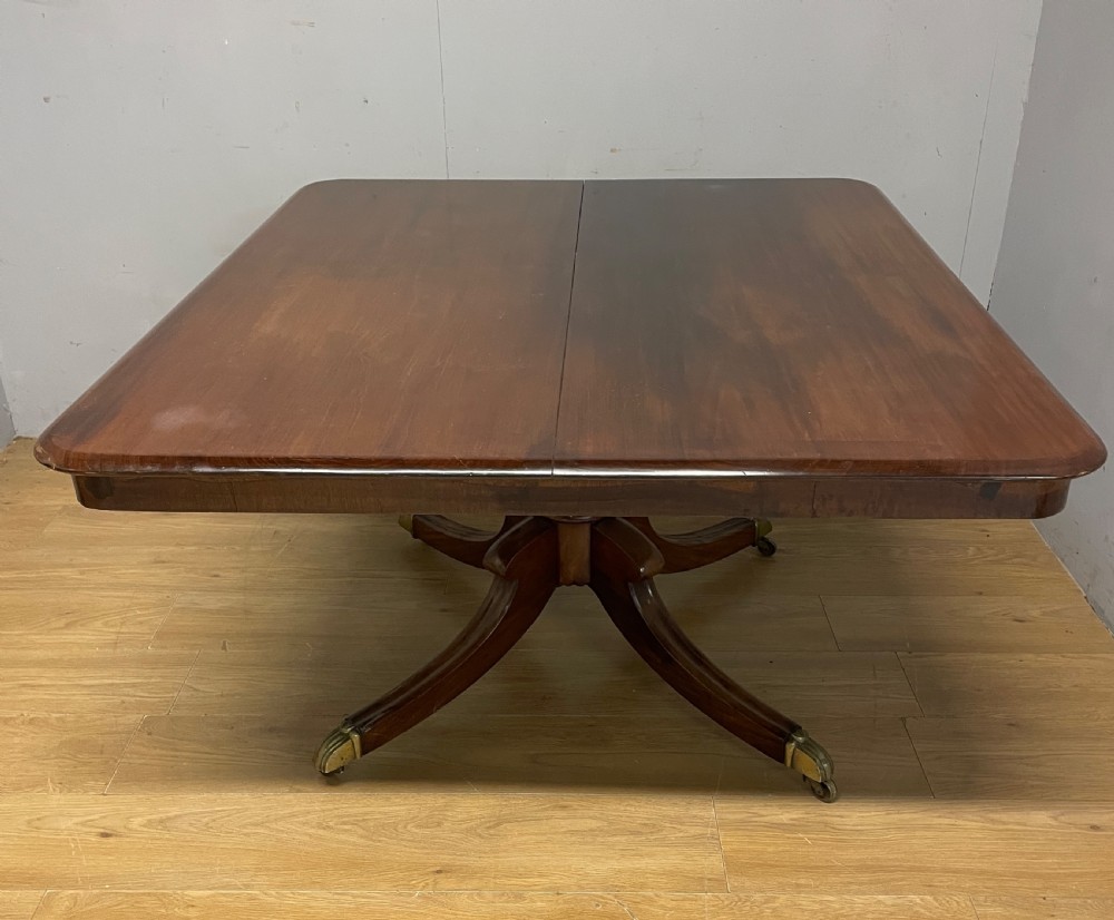 A William IV mahogany tilt-top dining table featuring a rectangular polished top with rounded corners supported by a central turned pedestal and four splayed legs with brass castors. View IV