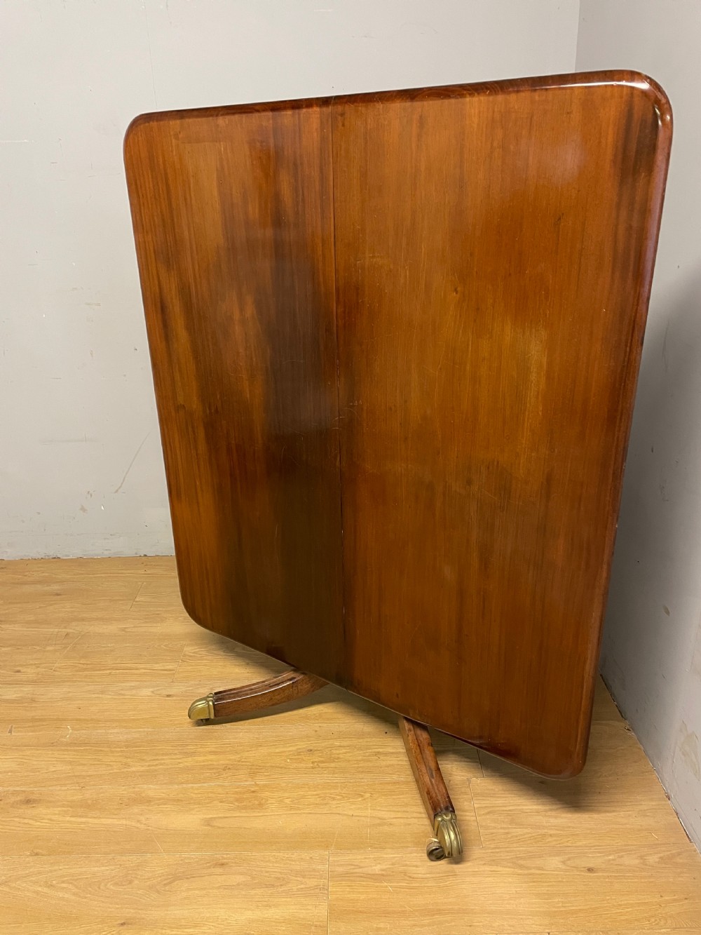 A William IV mahogany tilt-top dining table featuring a rectangular polished top with rounded corners supported by a central turned pedestal and four splayed legs with brass castors. View VII