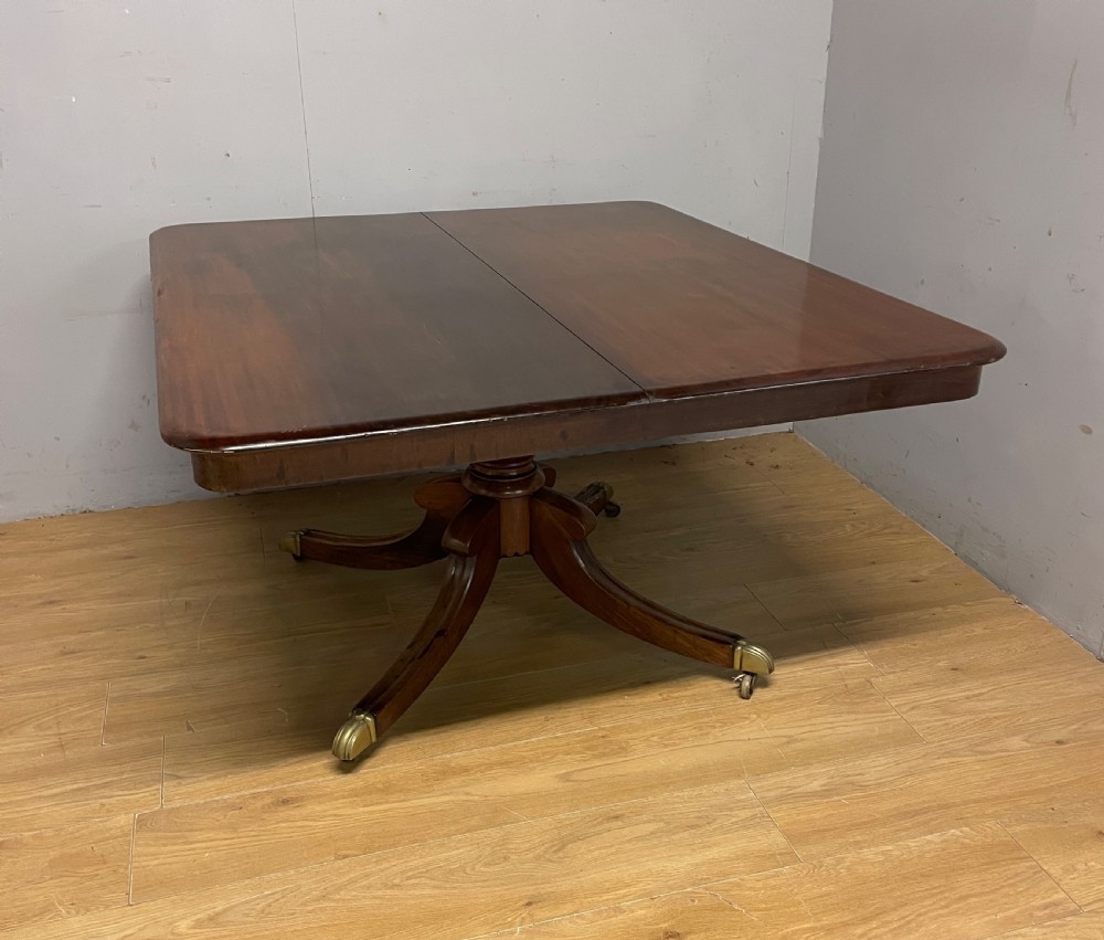A William IV mahogany tilt-top dining table featuring a rectangular polished top with rounded corners supported by a central turned pedestal and four splayed legs with brass castors. View IX