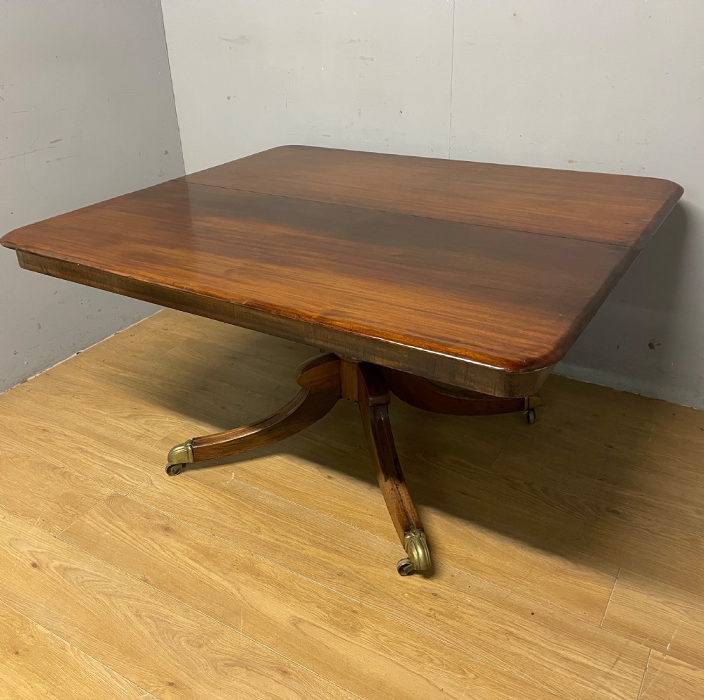 A William IV mahogany tilt-top dining table featuring a rectangular polished top with rounded corners supported by a central turned pedestal and four splayed legs with brass castors. View X