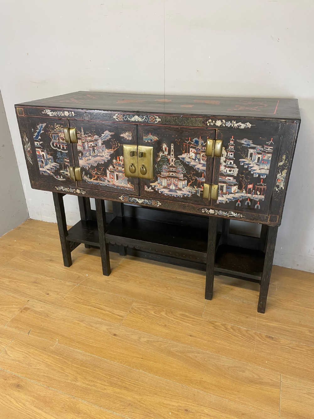 An antique black lacquered cabinet featuring hand-painted landscape scenes and large rectangular brass hinges, elevated on a dark wooden open stand.